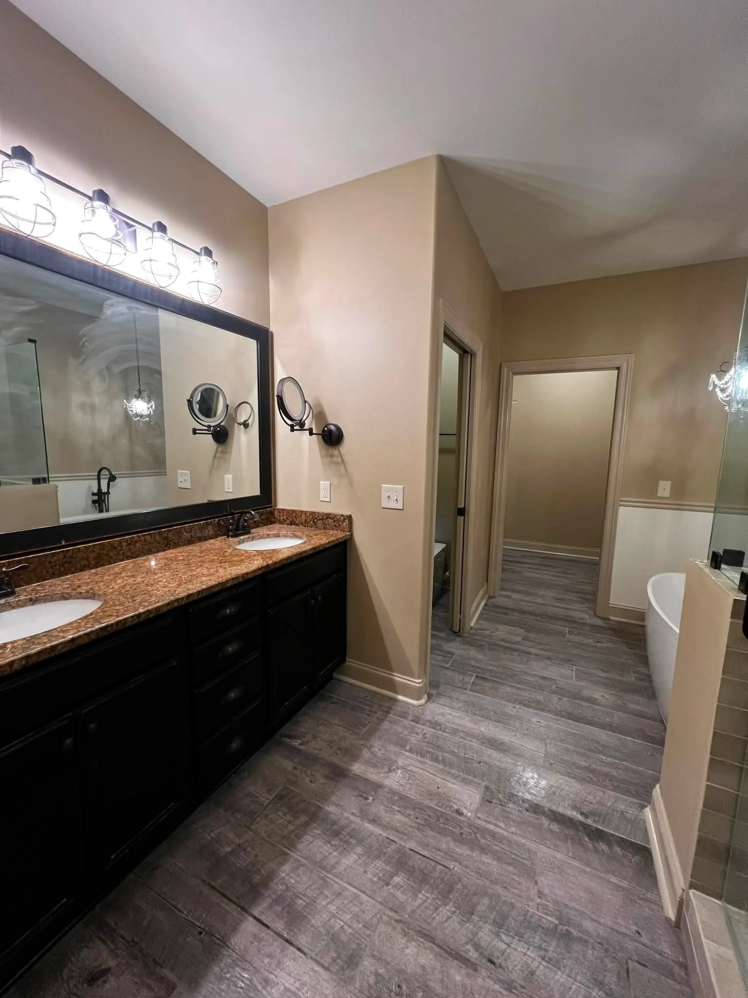 A bathroom with a double vanity sink with black cabinetry and granite countertops, large mirror, wall-mounted light fixture, wall-mounted mirrors, wood-look flooring, and a separate toilet area visible through an open door.