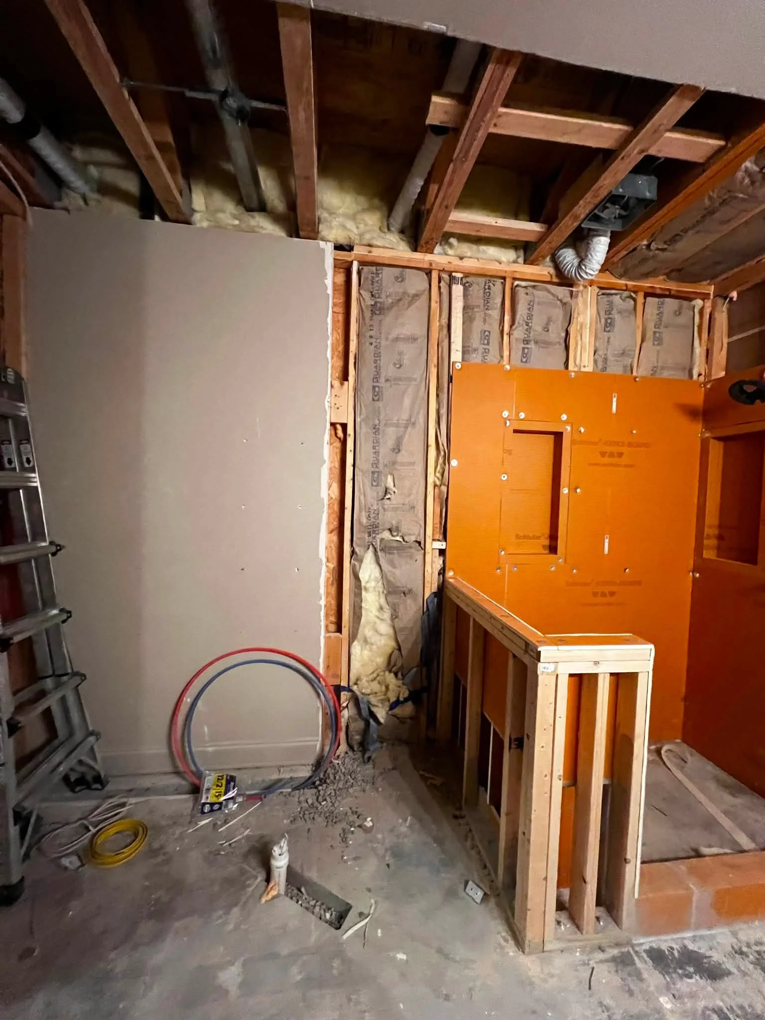 Construction area in a room with unfinished walls, insulation, and wooden framing. Building materials, tools, and electrical conduits are visible.
