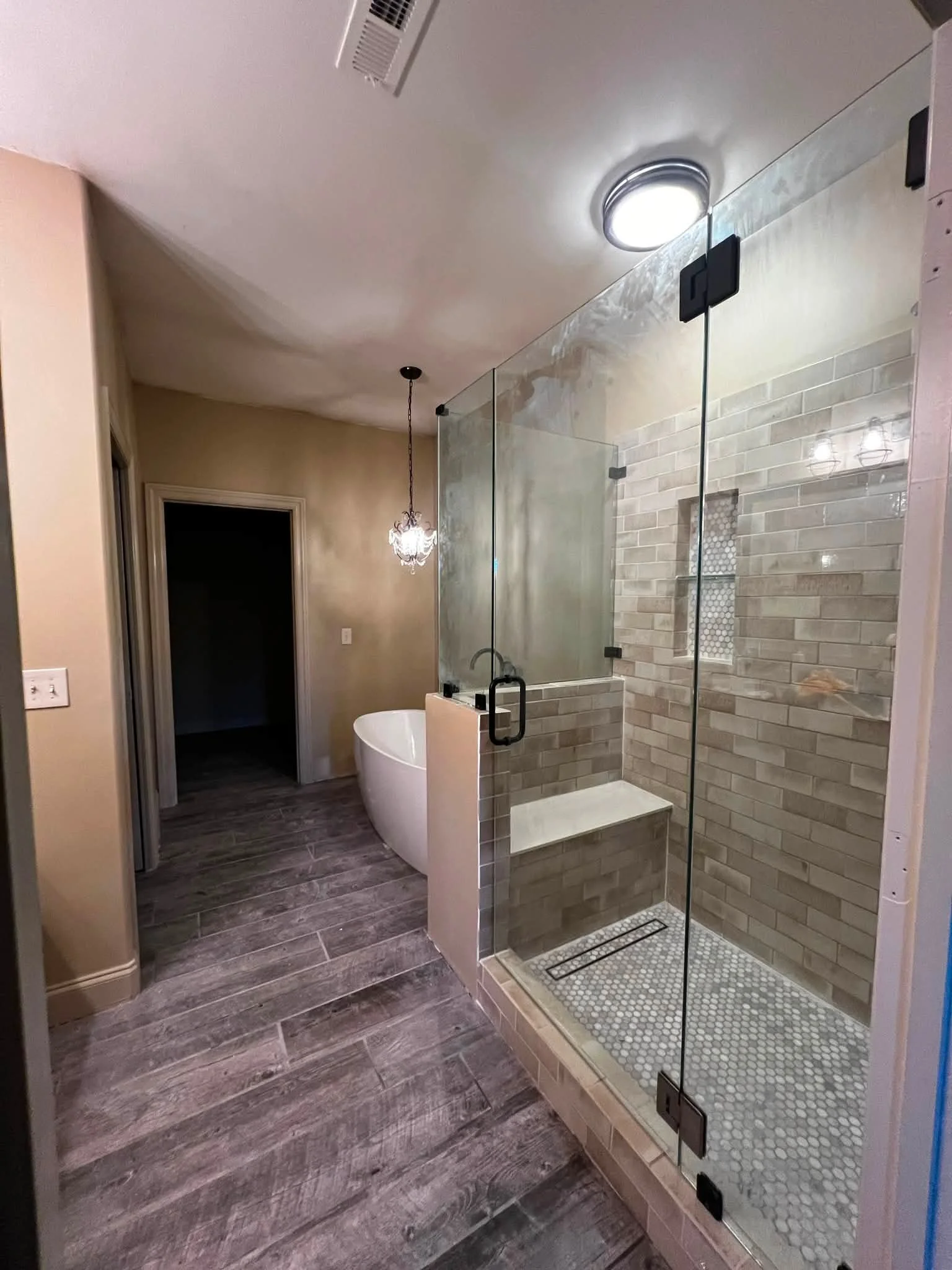 Bathroom with a walk-in glass shower, freestanding bathtub, chandelier light, wood-look tile flooring, and a doorway leading to a dark room.