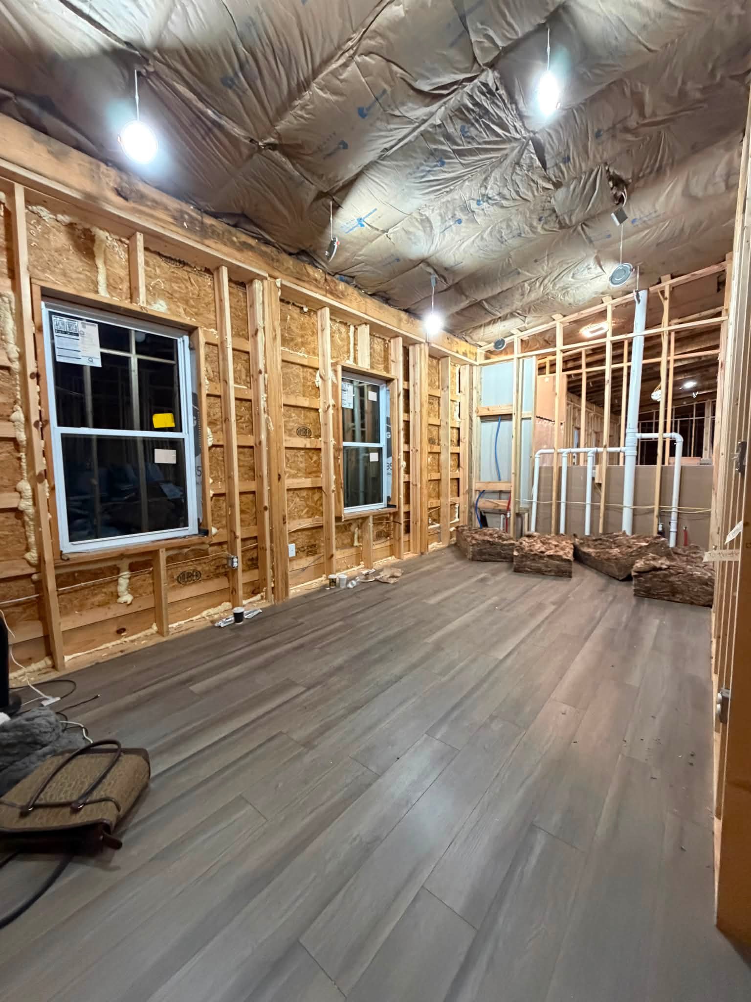 Interior of a house under construction with exposed wooden framing, insulation in the ceiling, two small windows, and unfinished walls. Wooden logs are on the floor, and construction tools and materials are visible.