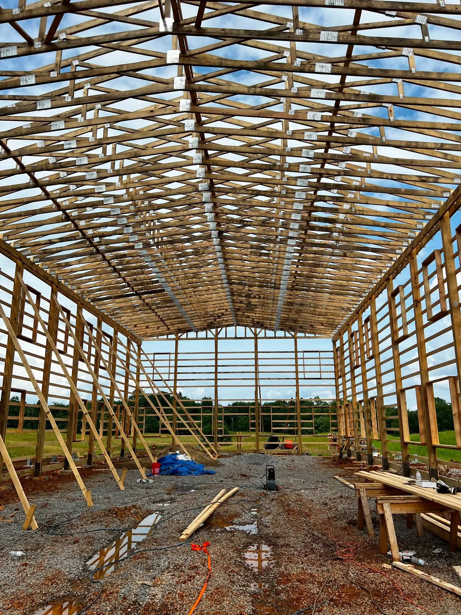 Looking into the wooden structure of a building under construction with a gabled roof, worker ladders, tools, scattered materials, and a wet gravel floor.