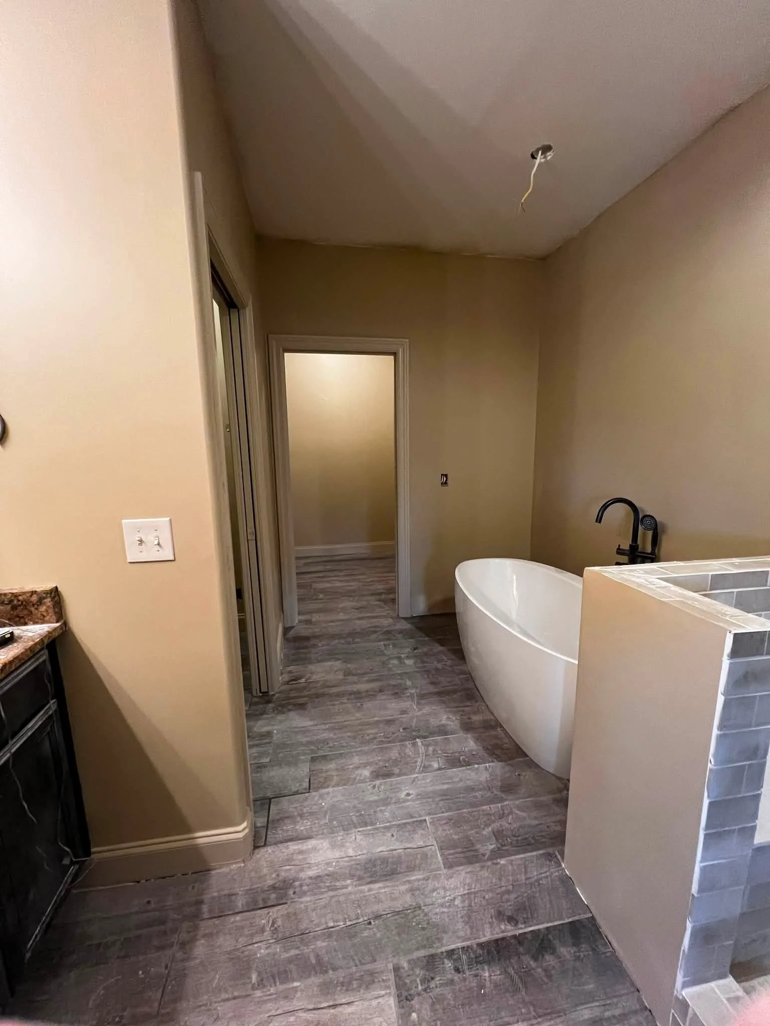 Empty bathroom with a freestanding bathtub, under-construction lighting fixture, gray wood-look flooring, beige walls, and a doorway leading to another room.