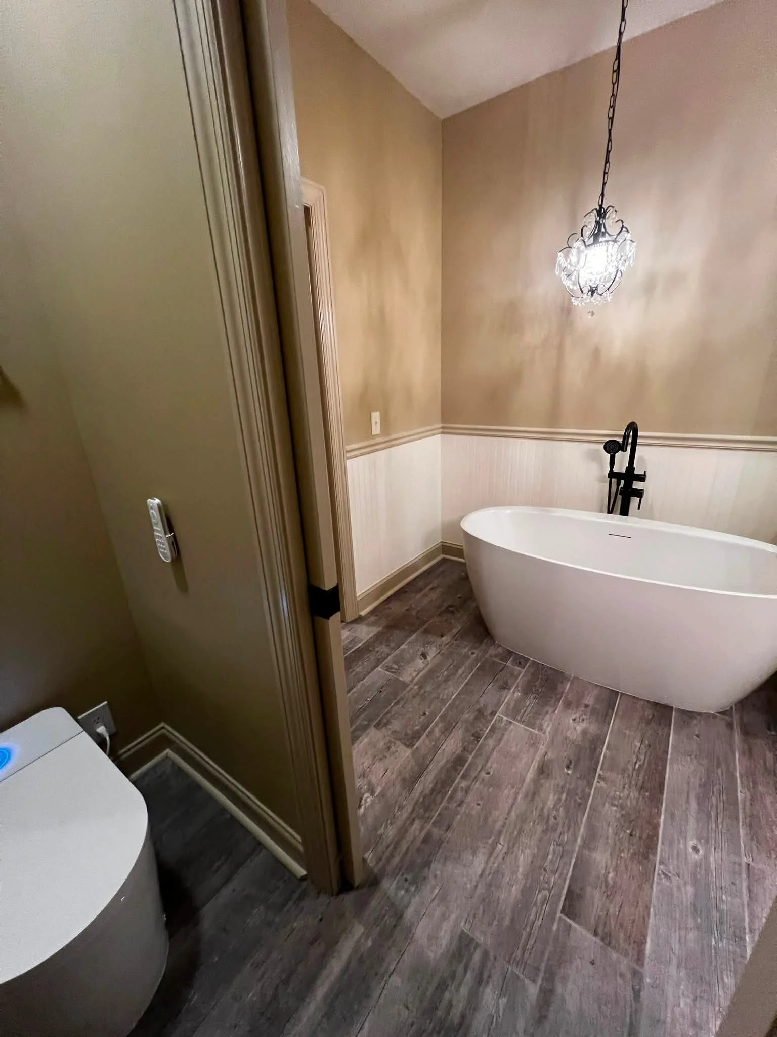 A bathroom with a modern bathtub, a black floor-mounted faucet, beige walls, wood-look flooring, a chandelier, and a partial view of a washing machine.