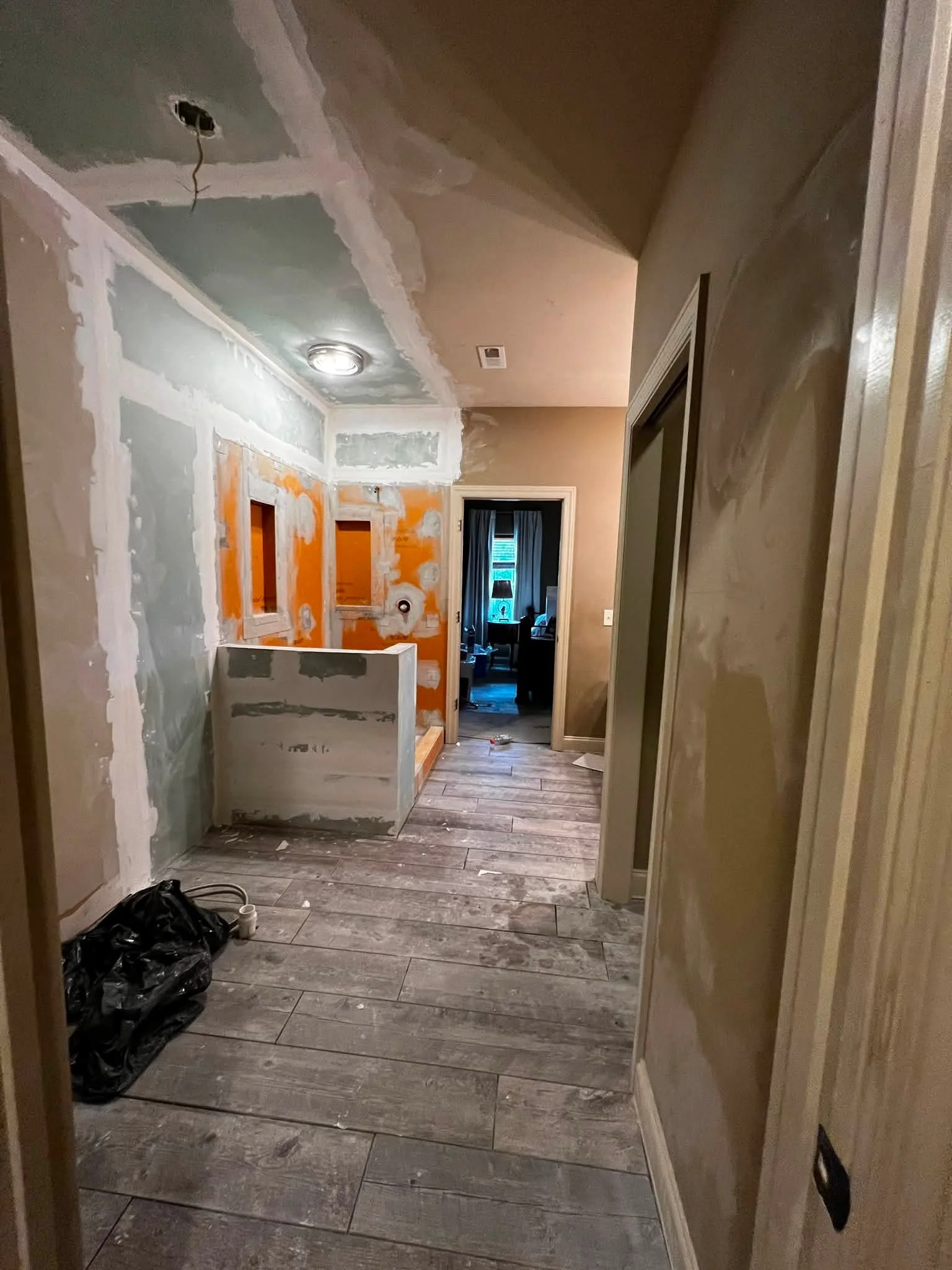 A hallway under renovation with drywall installation in progress, an open doorway revealing a room with curtains and a window, and construction materials and trash on the floor.