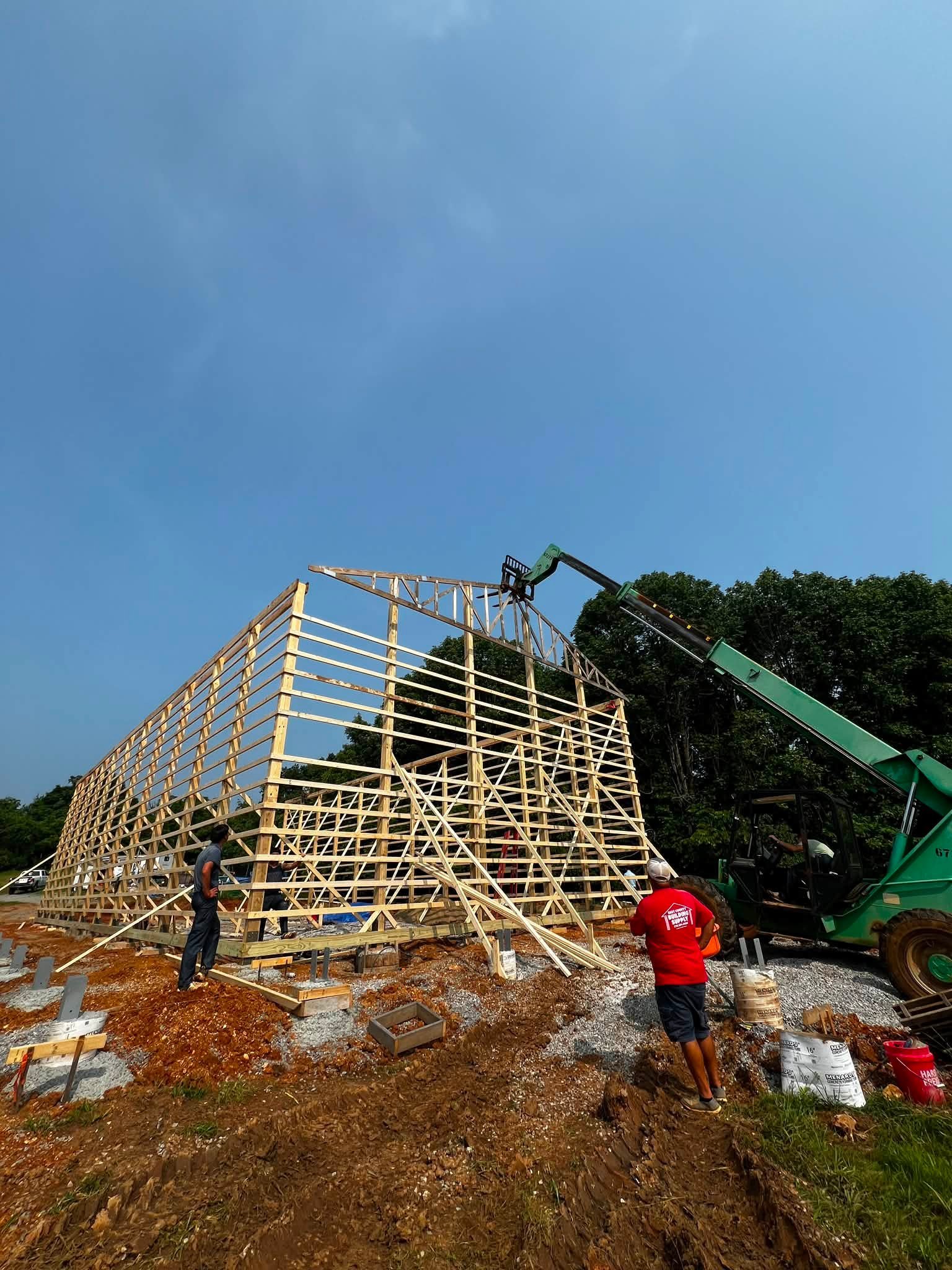 Wooden framing for a building under construction, with workers and a crane on a dirt site under a clear blue sky.