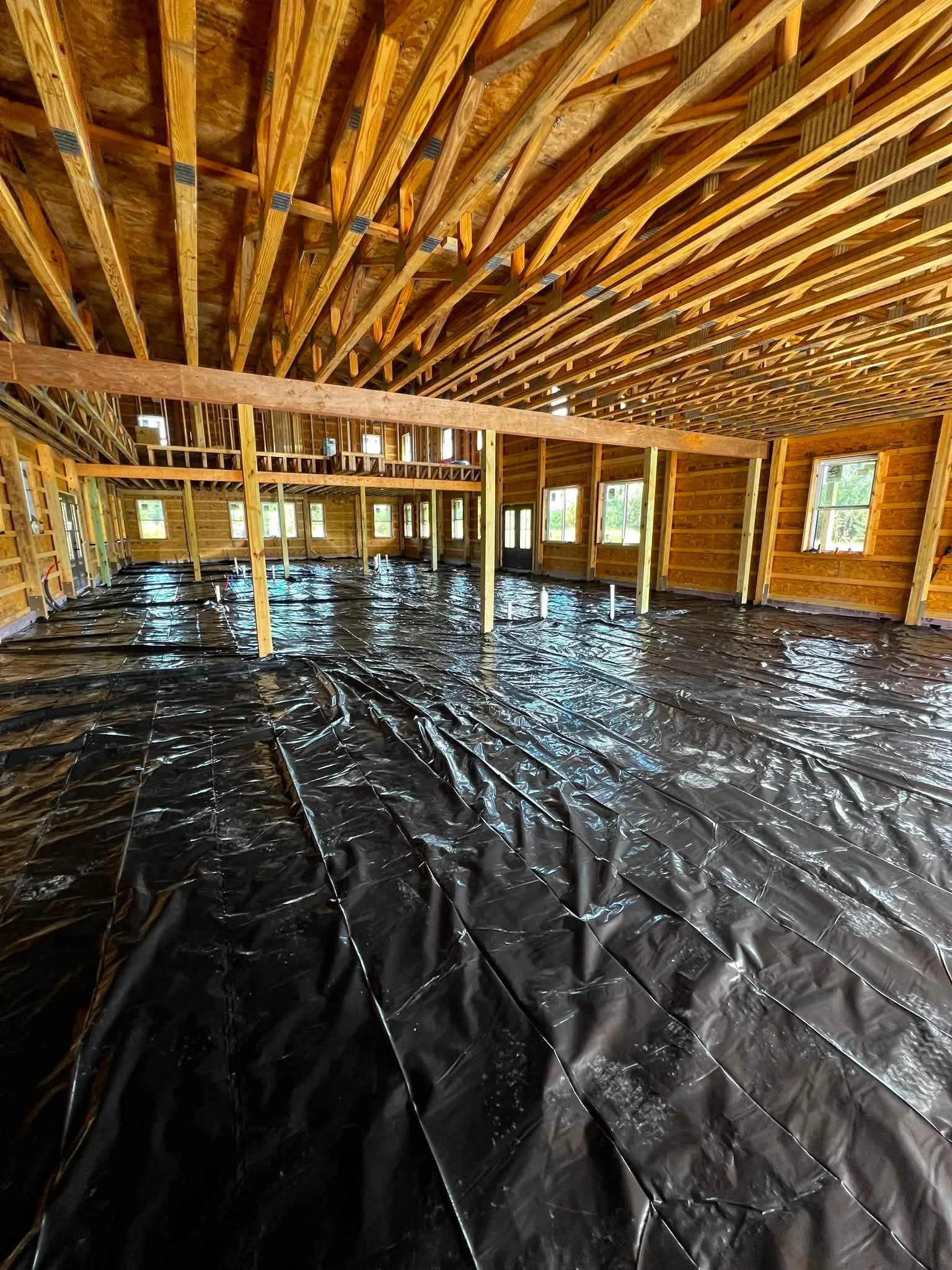 Interior of a house under construction with exposed wooden framing, black plastic sheeting on the floor, and multiple windows and doors.