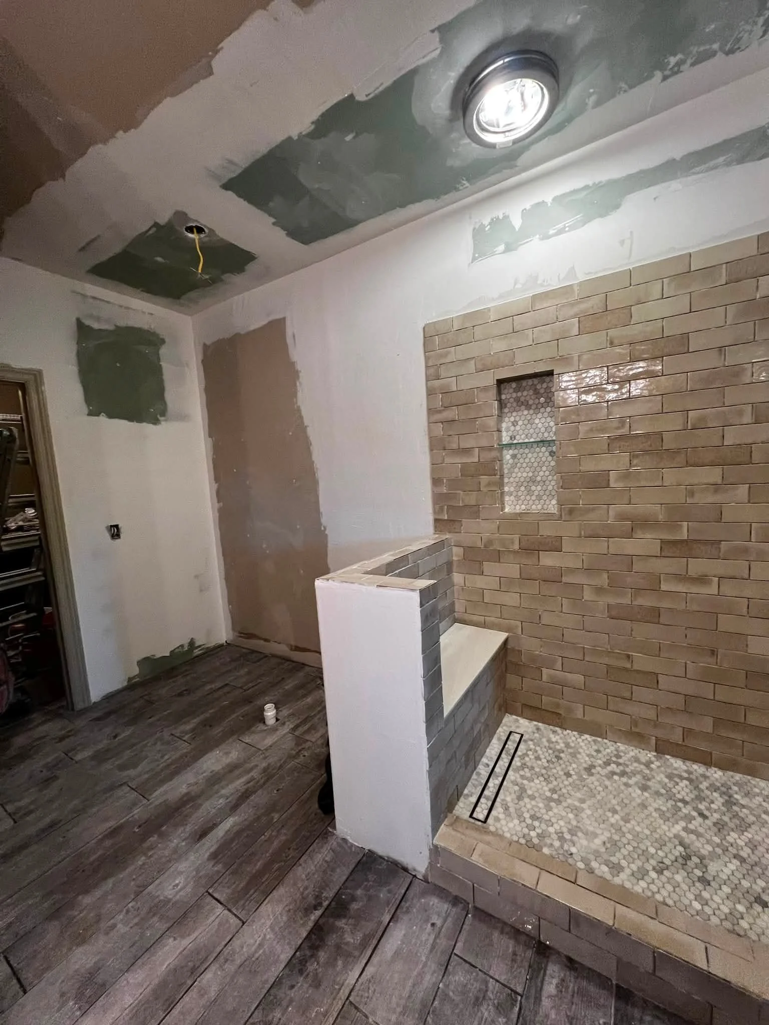 Interior of a room under renovation with an exposed ceiling with patches of drywall, a recessed light, a brick accent wall with a small niche, a small tile shower base with mosaic tiles, and dark wooden flooring.