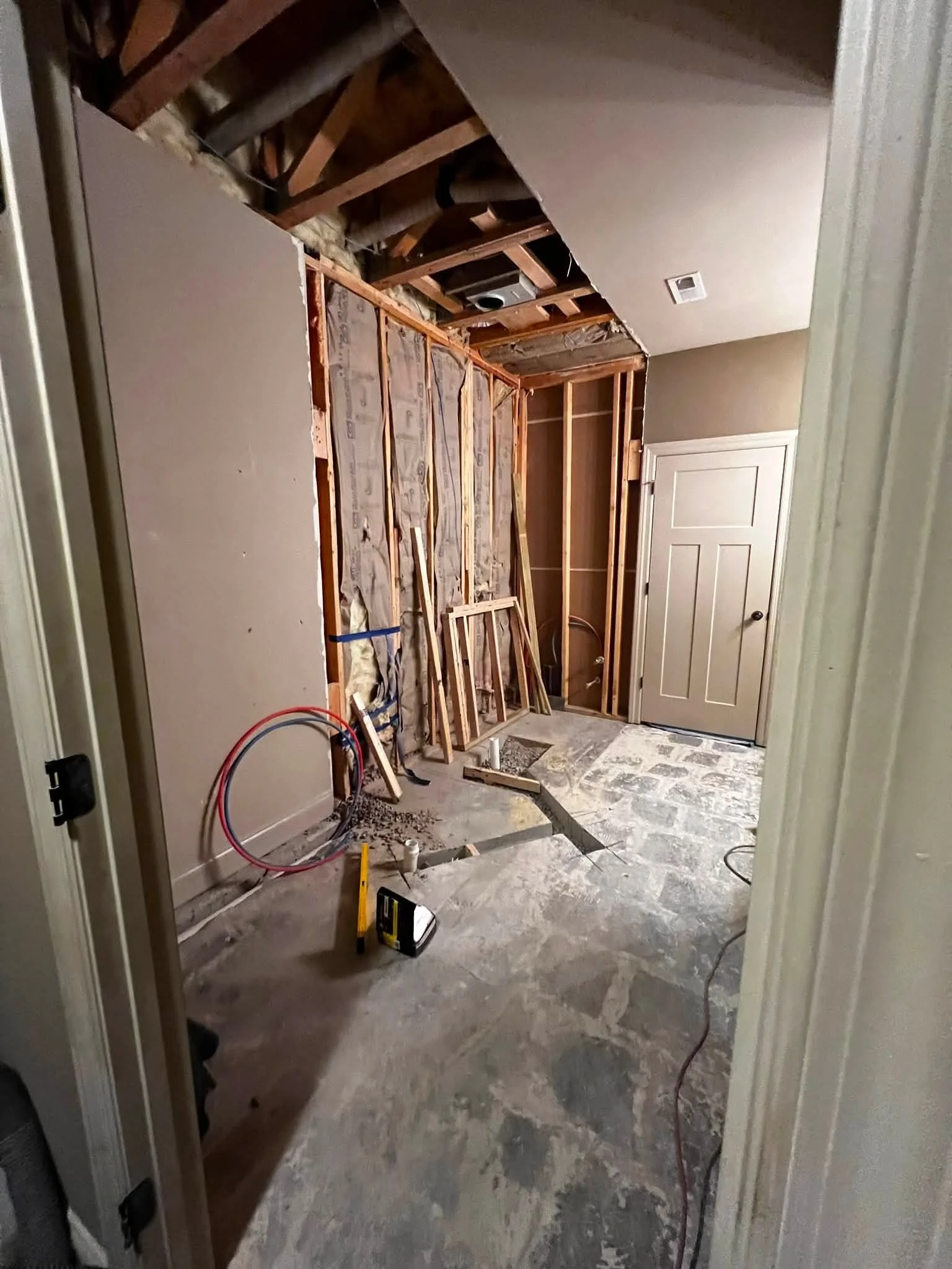 Room under renovation showing exposed wooden framing, electrical wiring, and partially torn drywall, with construction tools and materials on the floor.