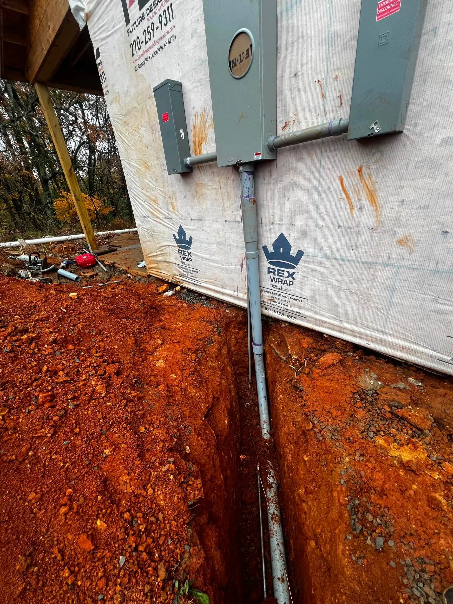A construction site with exposed electrical boxes and conduit on a house under renovation. The house wrap has branding and text on it. The ground is dug up with red soil and rocks. There is some scattered debris and tools, and trees with autumn folia