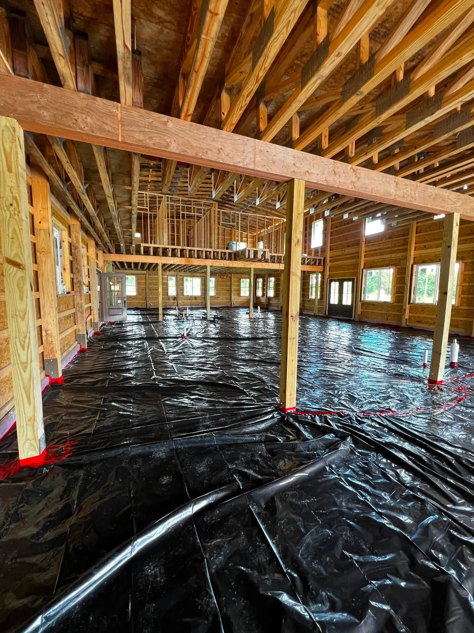 Inside of a house under construction with exposed wooden framing, black plastic vapor barrier on the floor, and multiple windows.