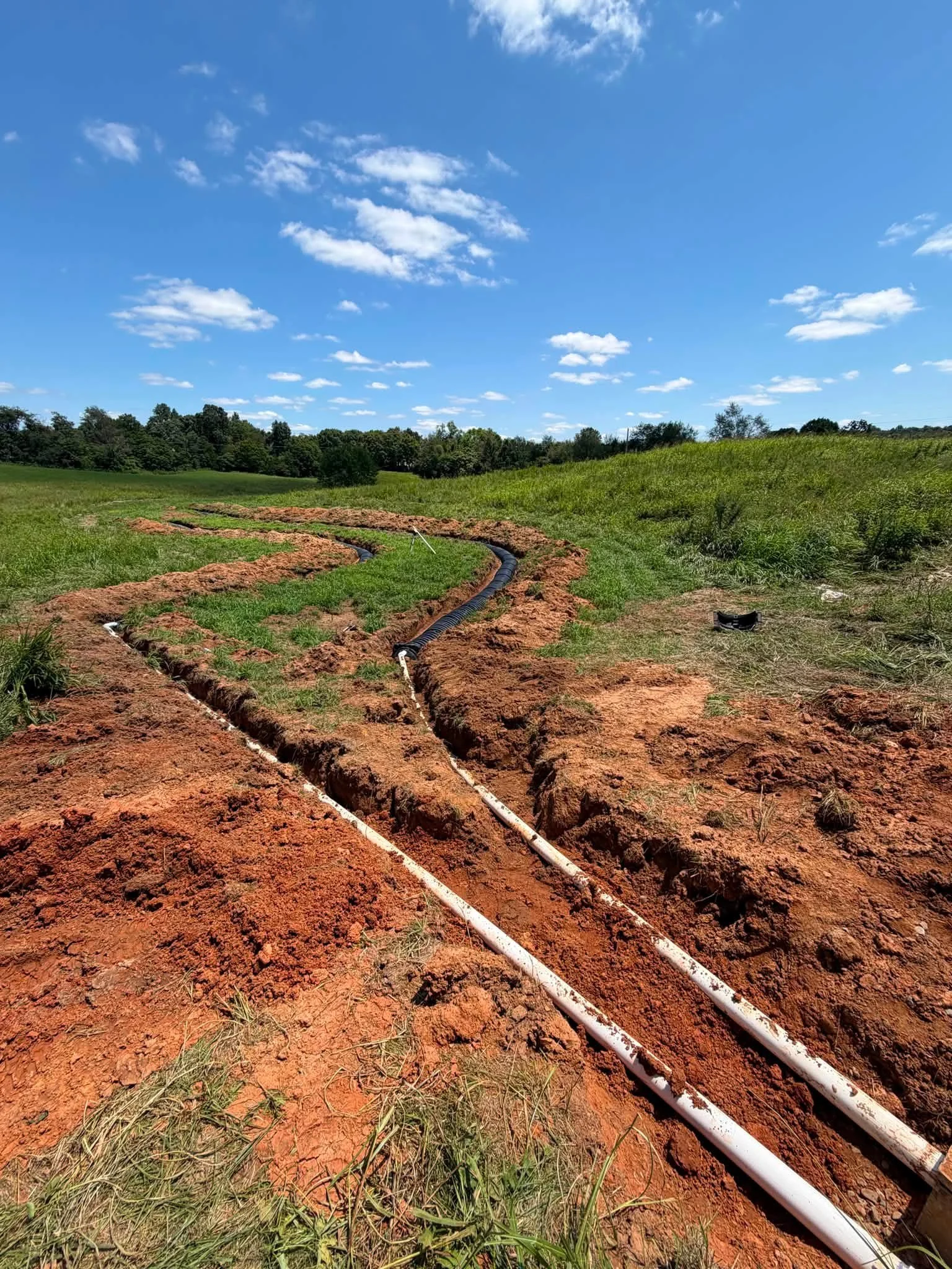 A rural landscape with a bright blue sky and scattered white clouds, green grassy fields with a gentle hill, and two white pipes laid in the ground, likely part of an irrigation system.