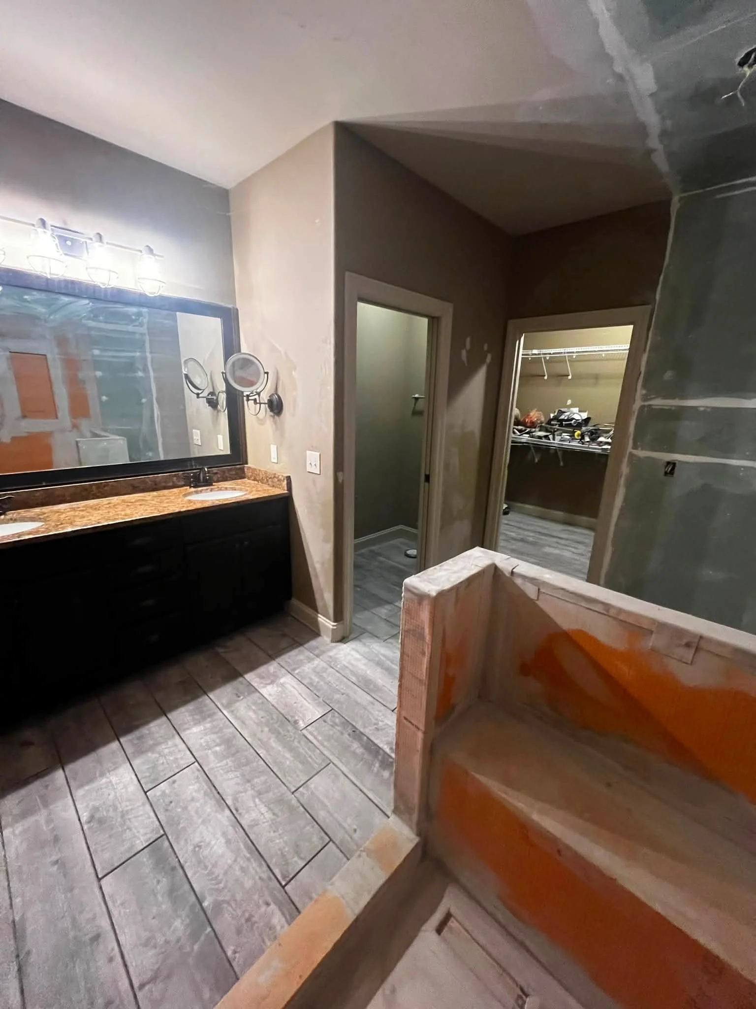 A bathroom under renovation with a double vanity with granite countertop, wall-mounted mirror, and two faucet sinks. There is construction debris, an unfinished wall with drywall, and an open closet in the background.