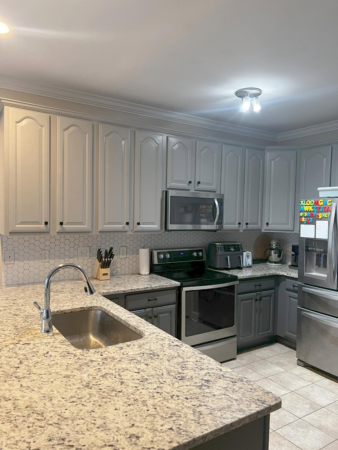 Kitchen with white cabinets, granite countertops, stainless steel appliances including a microwave, oven, and refrigerator. Kitchen tools on the counter, a sink with a faucet, and decorative hexagon tiles backsplash.