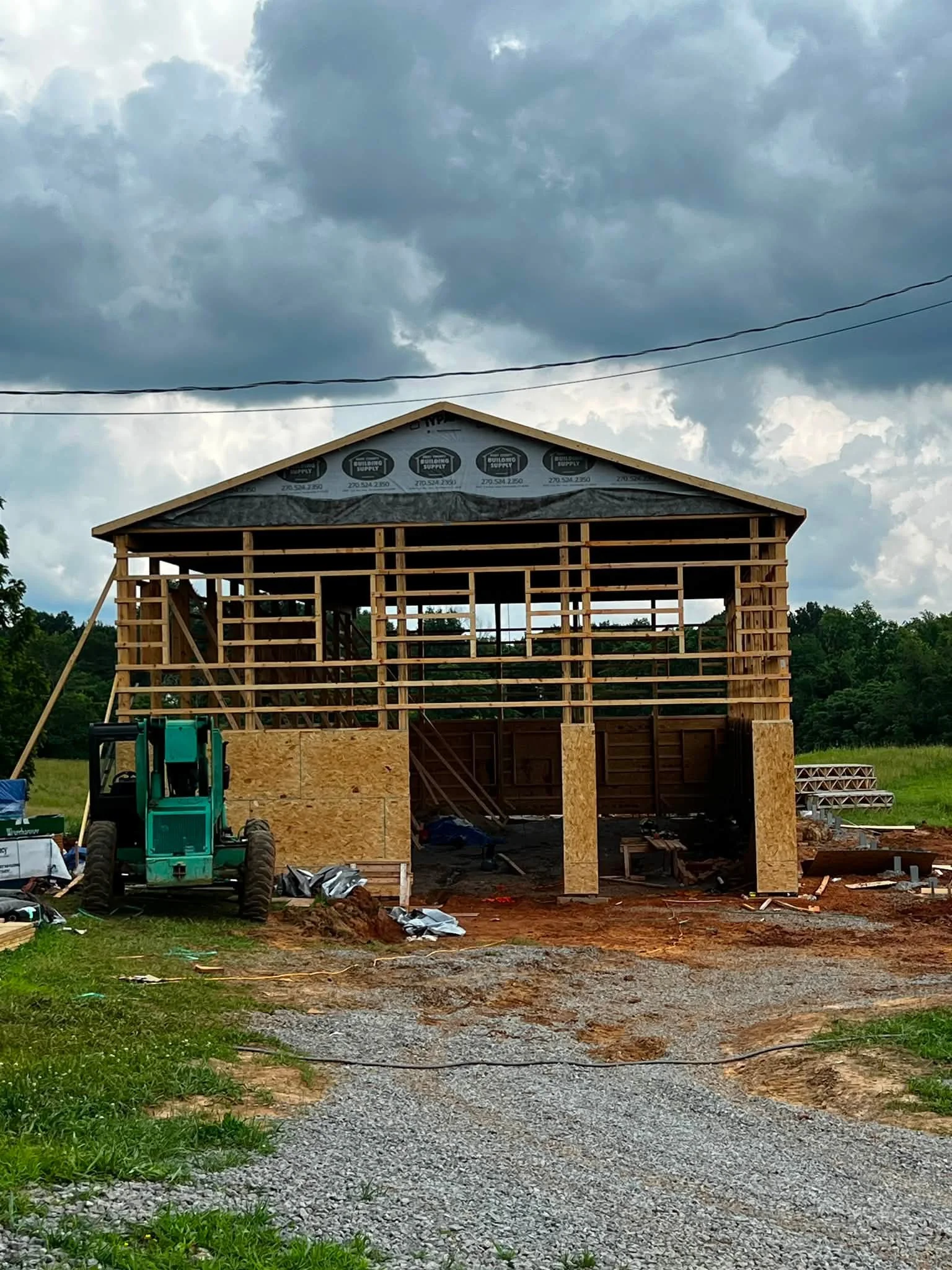 A house under construction with wooden framing and a partially installed roof, set against a cloudy sky with trees in the background.