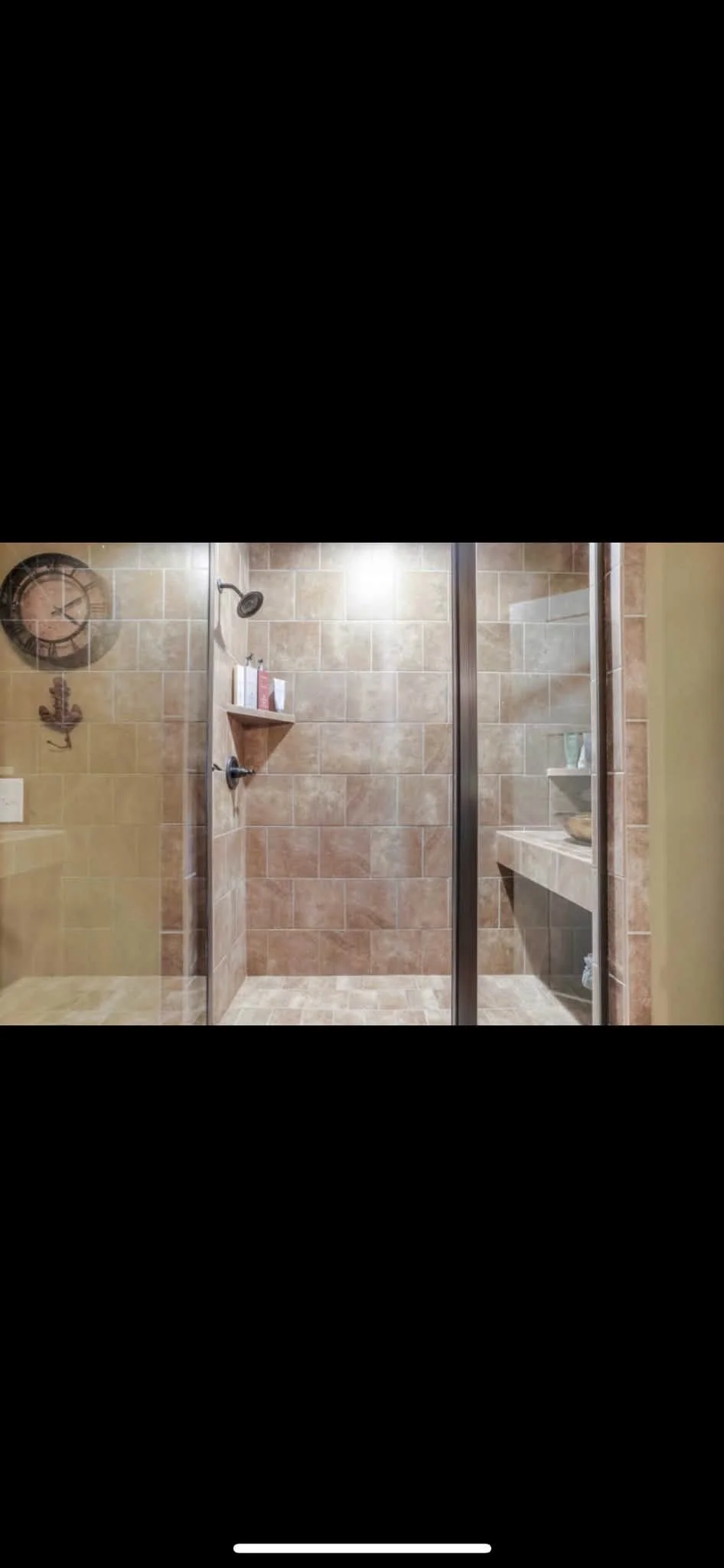 Bathroom shower area with brown tiled walls and floor, glass enclosure, wall-mounted shower head, a small corner shelf with toiletries, and a built-in countertop with a bowl and toiletries.