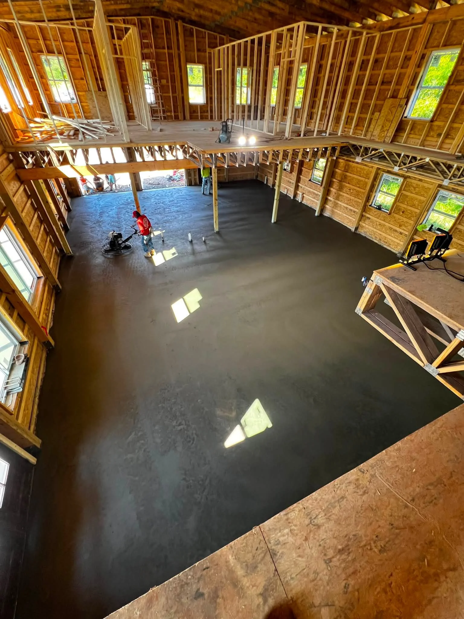 Interior of a house under construction with exposed wooden framing, windows, and a concrete floor where workers are smoothing the surface.