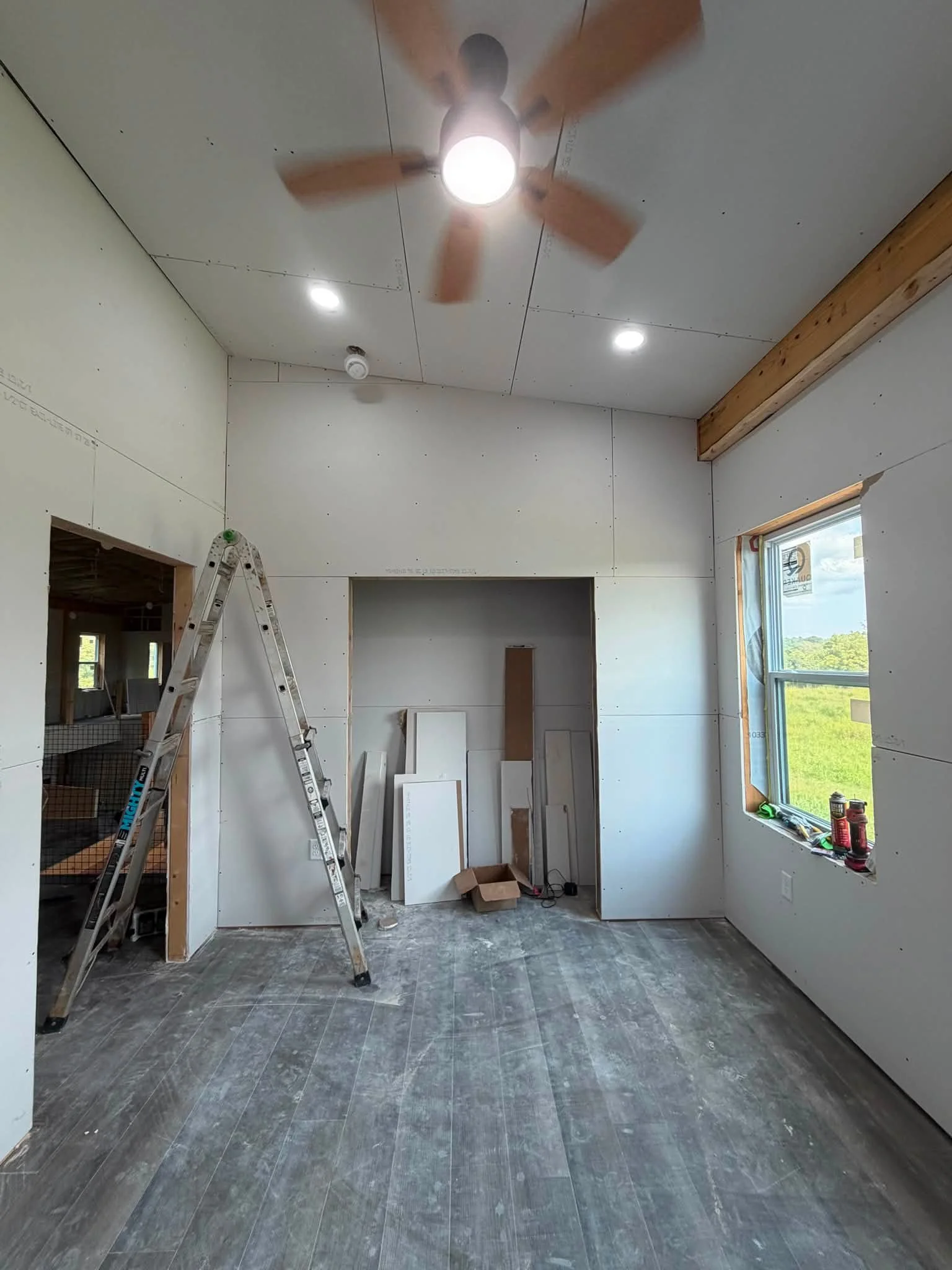 Interior of a room under construction with drywall panels, a ladder, and construction tools, featuring a window and ceiling fan.