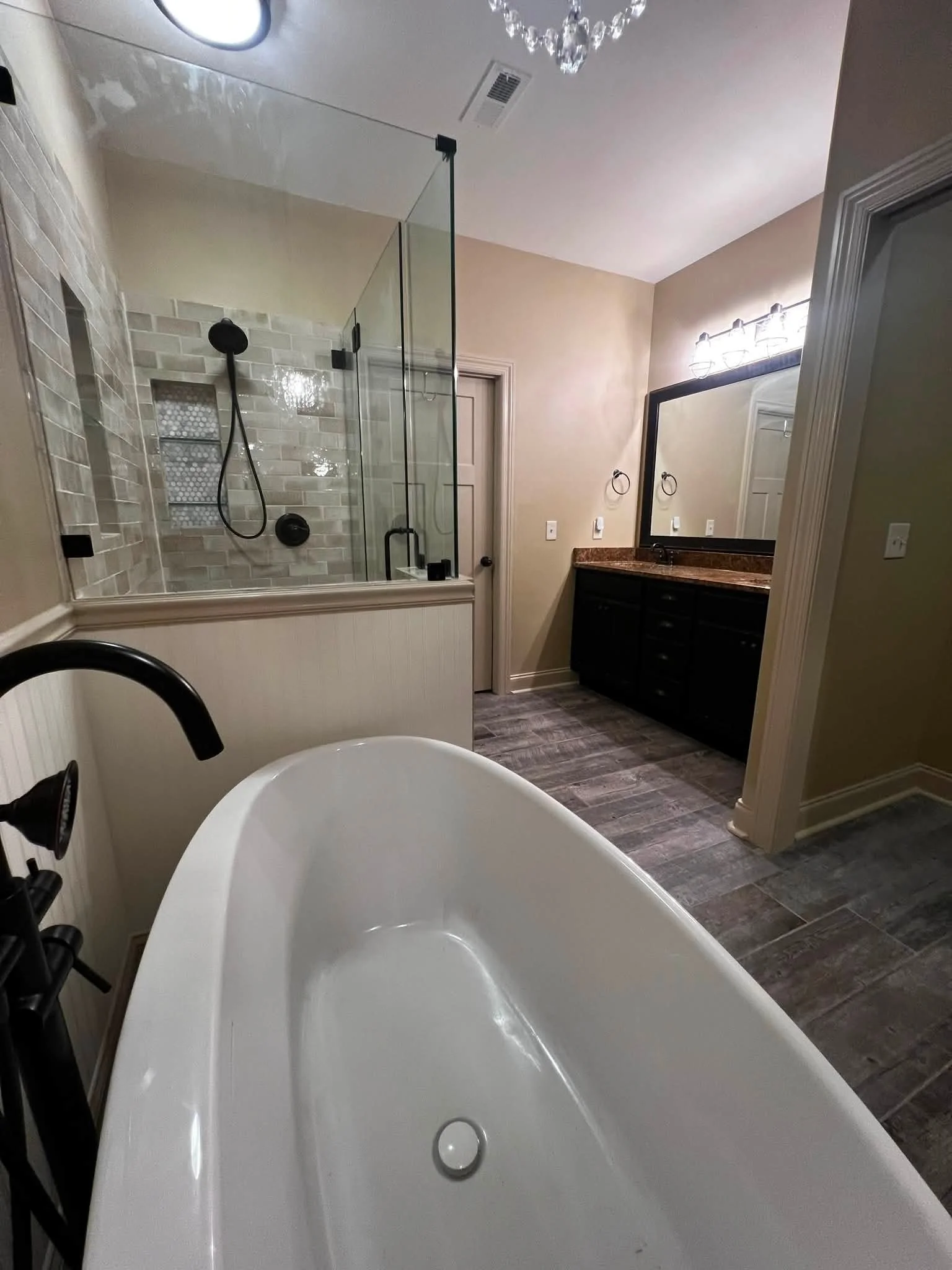 A bathroom with a white freestanding bathtub, black fixtures, a glass shower enclosure with beige tile inside, a mirror with vanity lighting, dark wood vanity cabinet, beige walls, and wood-look tile flooring.