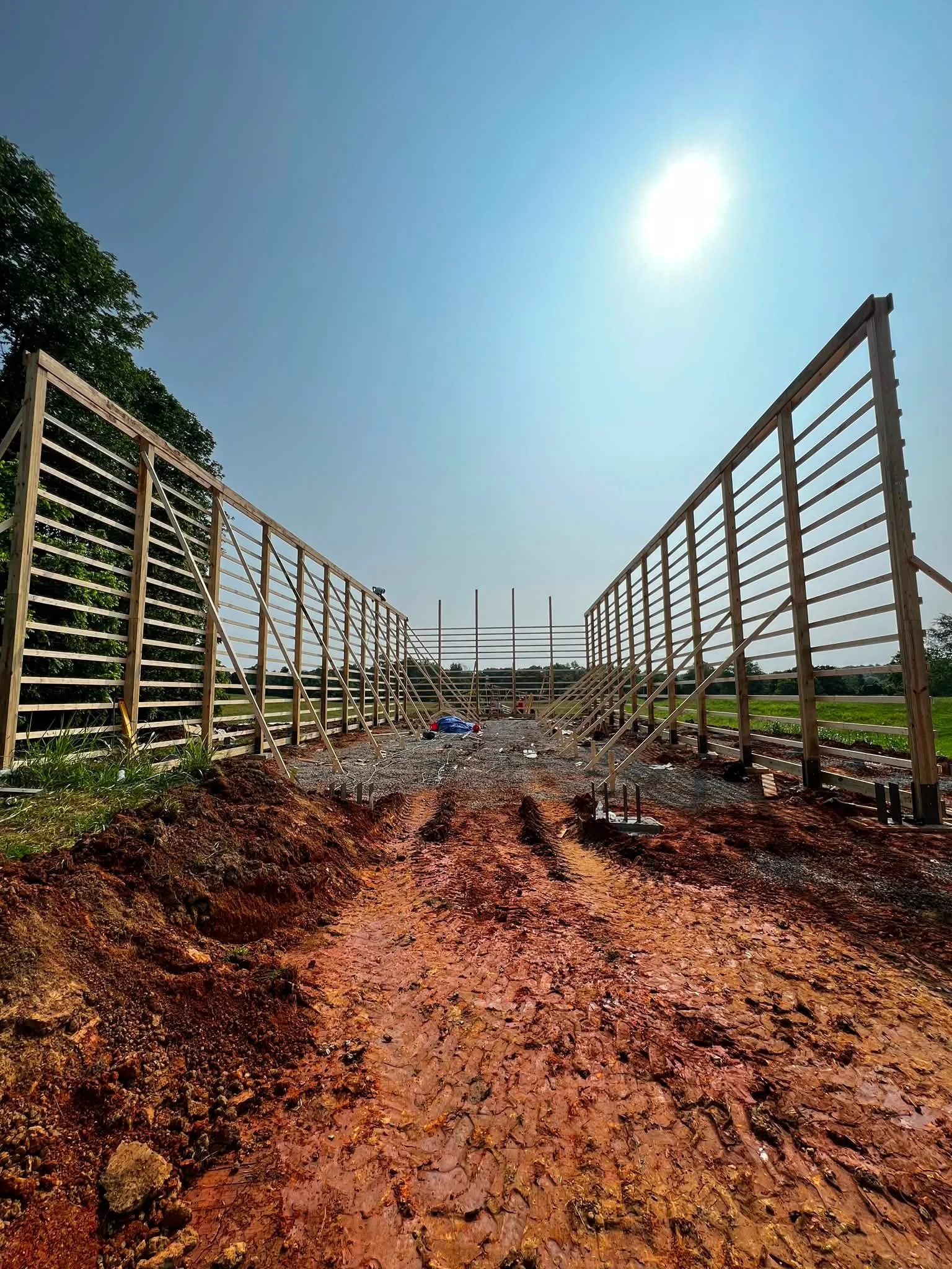 Construction site with unfinished wooden framing, dirt ground with tire tracks, and clear blue sky with the sun overhead.