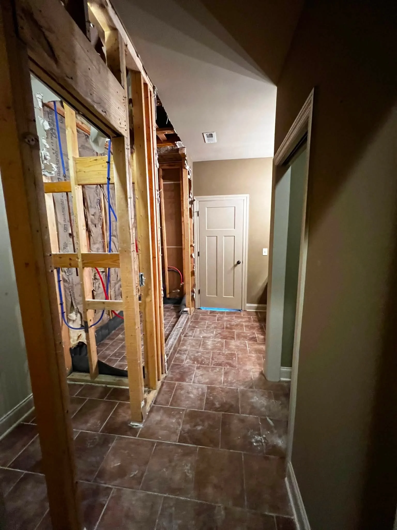 Interior of a room undergoing renovation with exposed wooden framing, partially finished walls, a closed door, and brown tiled flooring.