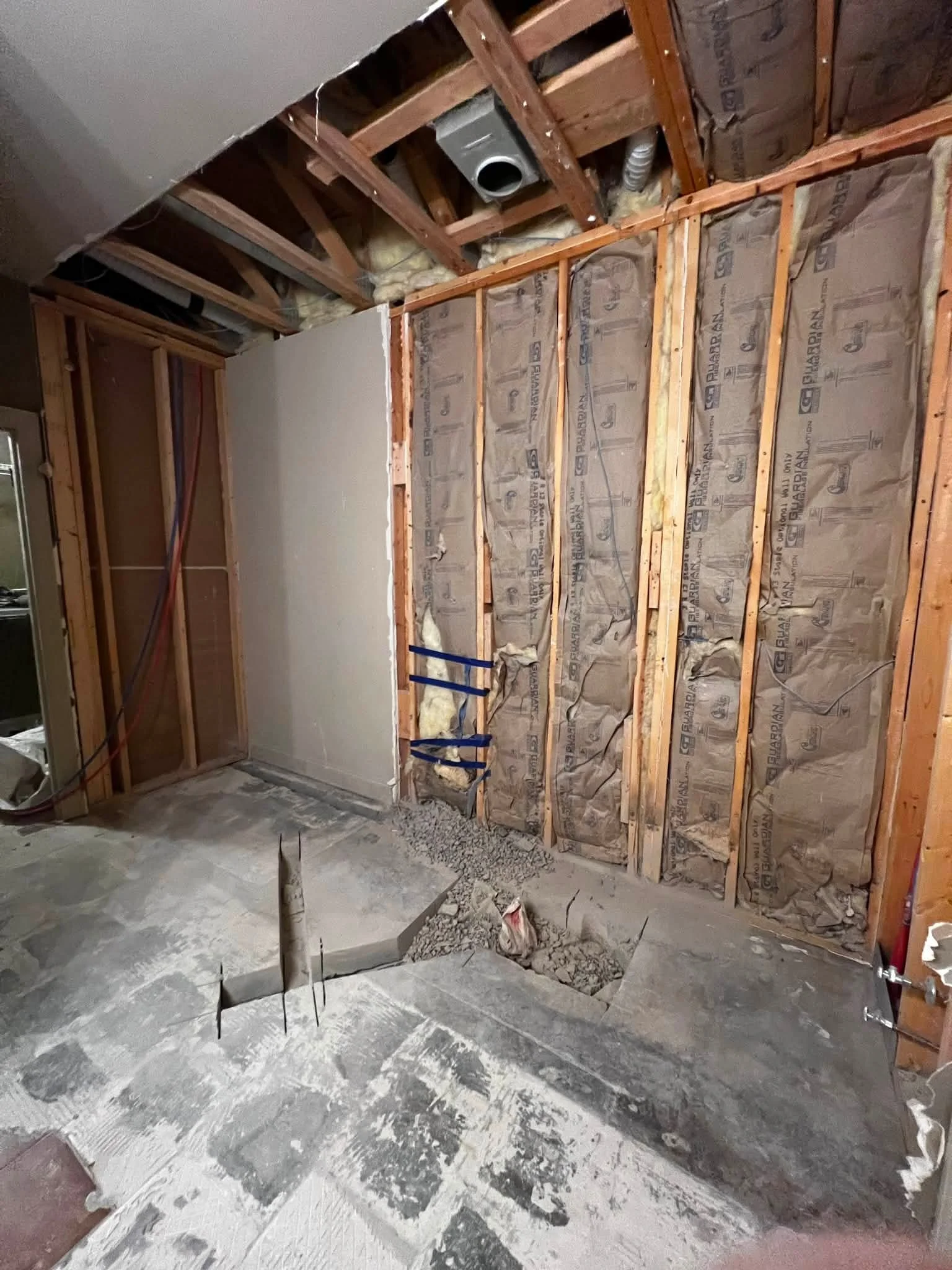 Drywall partially removed from a wall, revealing insulation, wooden studs, and ductwork in a construction or renovation site. The floor has construction debris and dust.