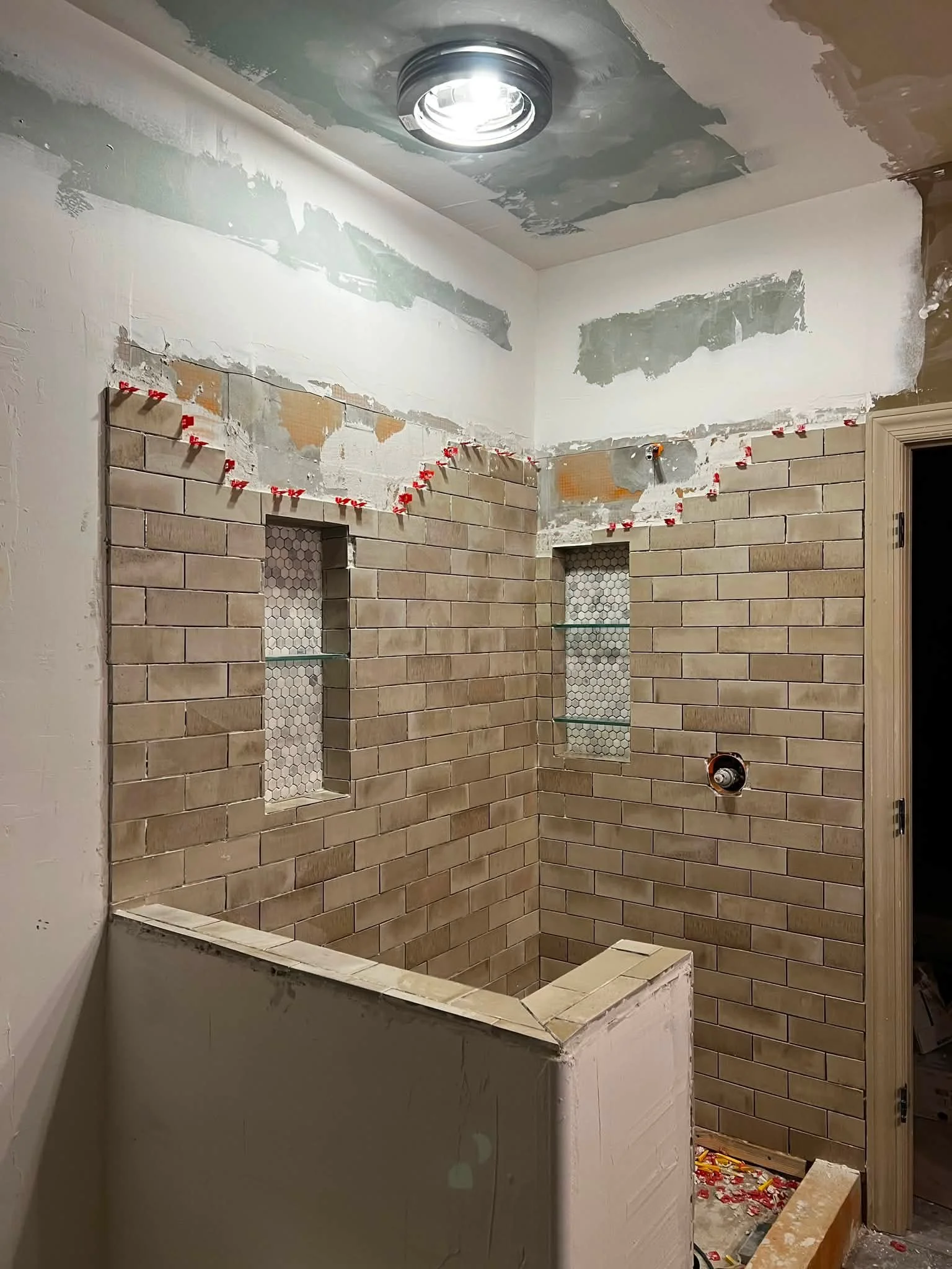 Bathroom under renovation with beige subway tiles on the wall, two built-in niches with honeycomb tiles, construction tools, and a temporary wall blocking part of the space.