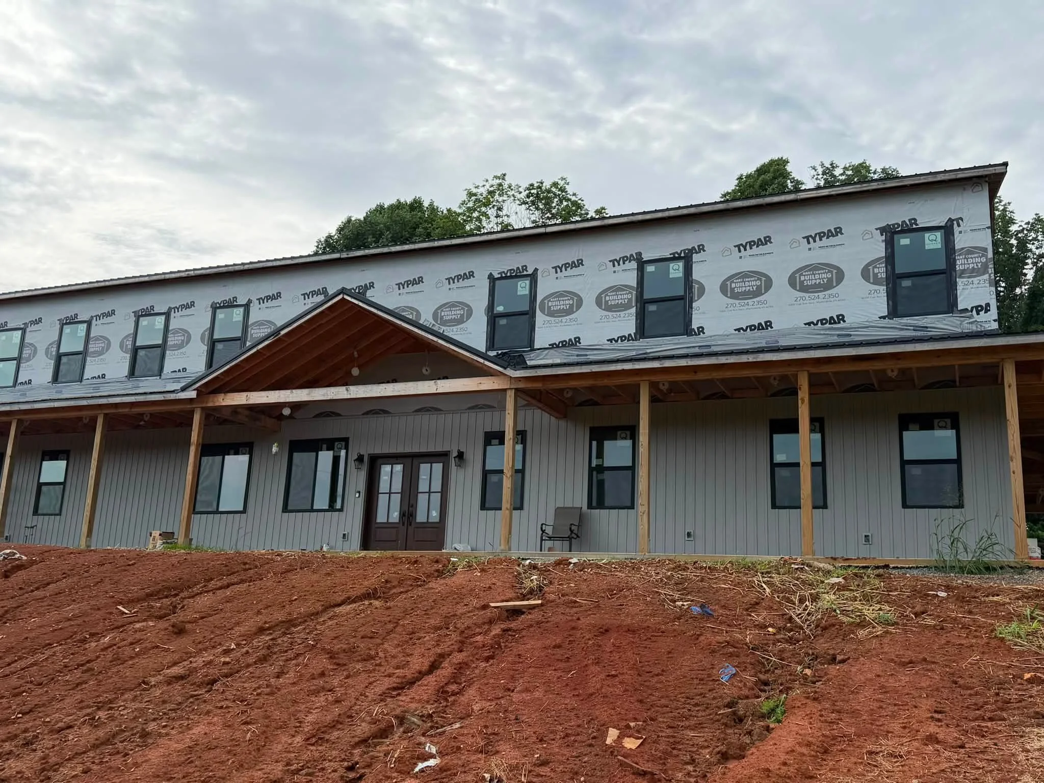 The image shows a house under construction, with the exterior finished but the roof not yet complete. There are multiple windows installed, and some parts of the house are covered with Typar building wrap. The foreground has reddish-brown dirt, and there is some greenery and trees in the background under a cloudy sky.