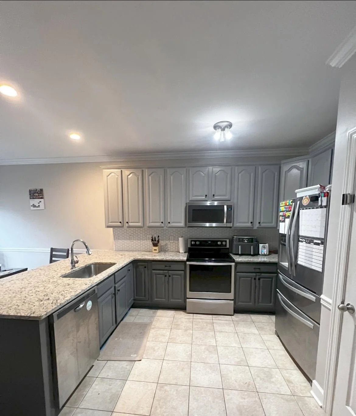 Kitchen with gray cabinets, granite countertops, stainless steel appliances including a microwave, oven, and refrigerator, tiled floor, and a small beige rug near the sink.