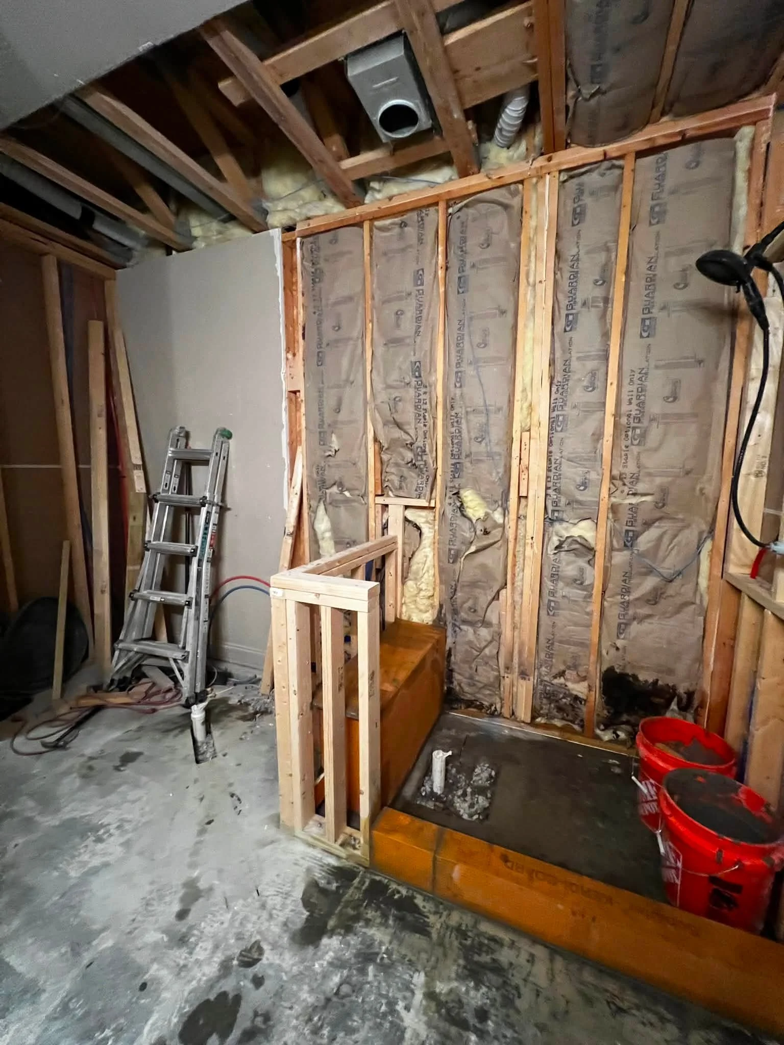 In this photo, there is a partially constructed or renovated room with exposed wooden wall framing, insulation, and partially installed drywall. There is a silver ladder, a red bucket, and some construction debris on the concrete floor.