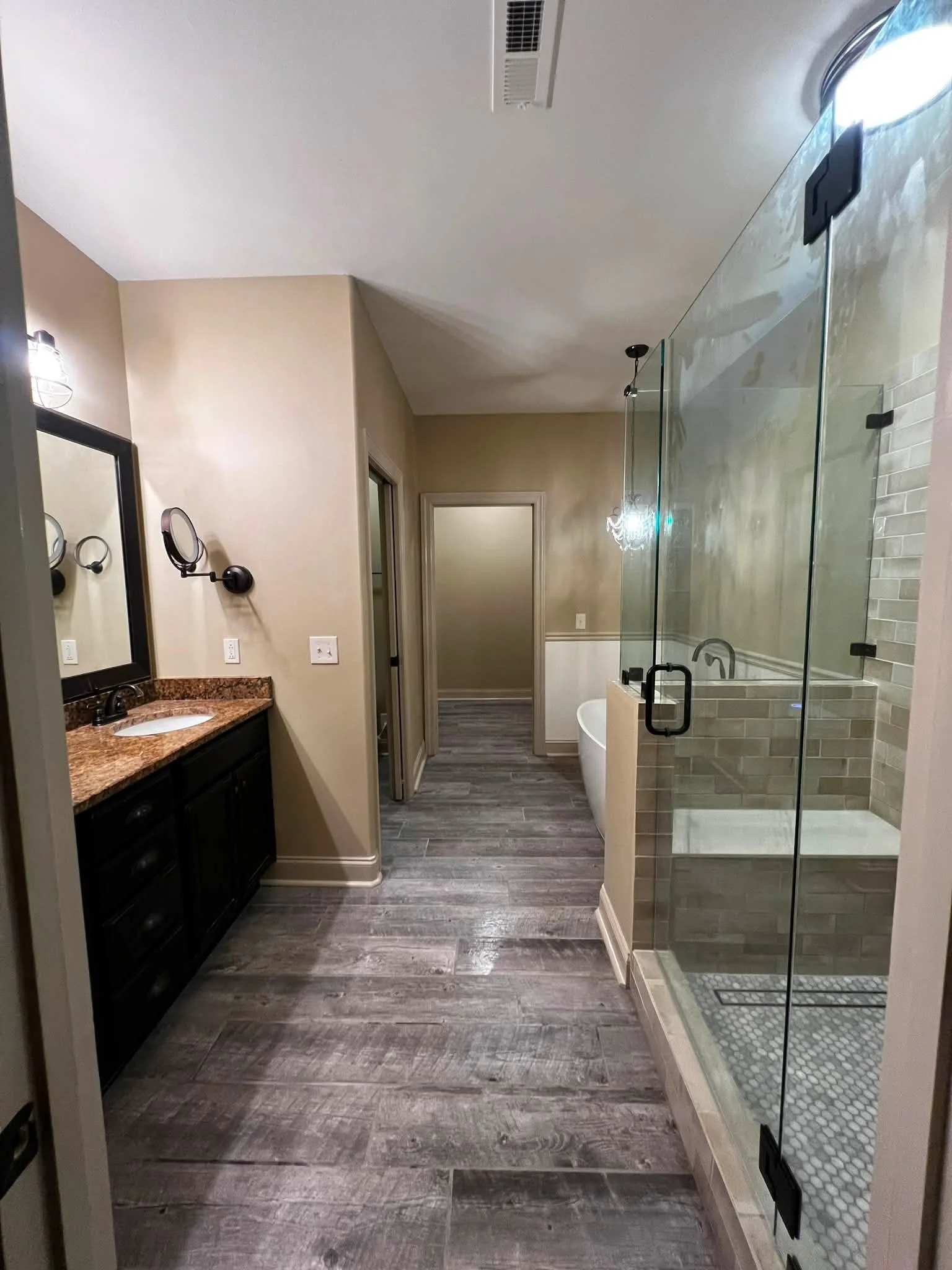 Bathroom with beige walls, dark wood vanity, granite countertop, two wall-mounted mirrors, two light fixtures, glass shower enclosure, white free-standing bathtub, wooden flooring.
