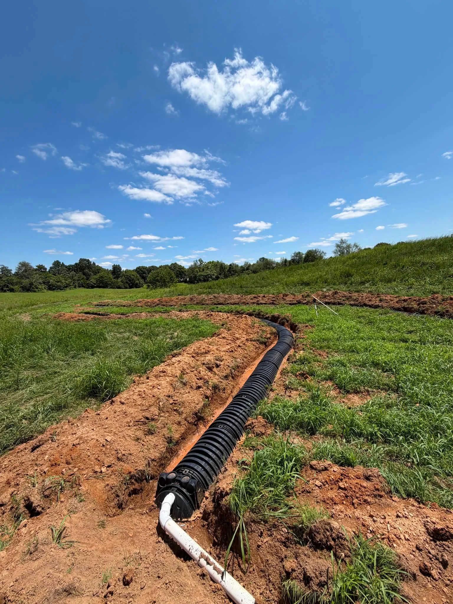 Installing underground piping in a grassy field under a blue sky with scattered clouds.