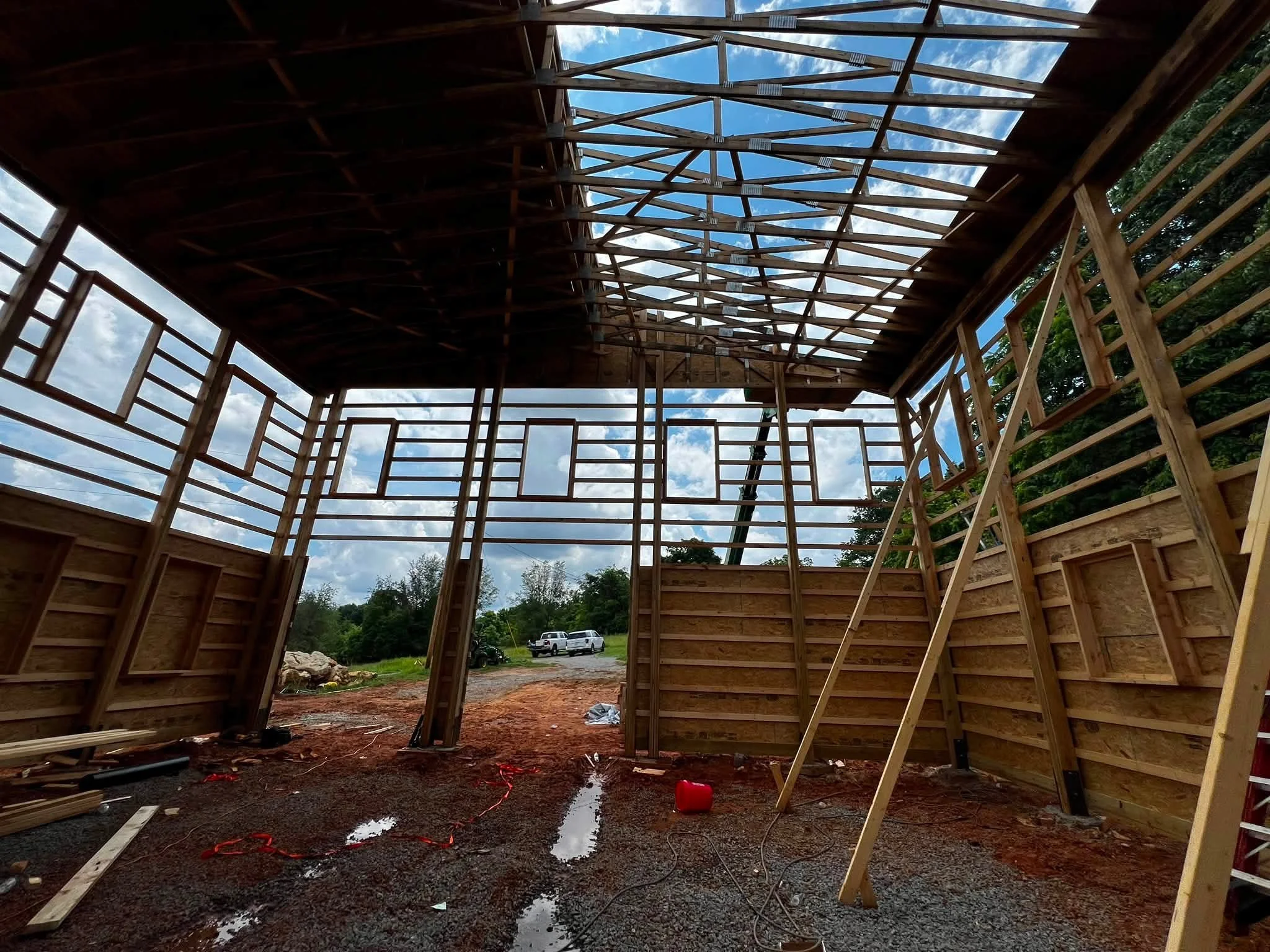 Construction site of a wooden building with a partially completed roof, showing blue sky and clouds through open roof structure and trees outside.