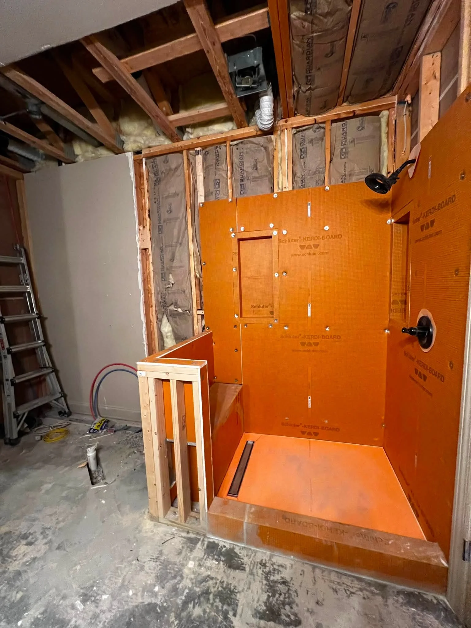 Construction site of an elevator with orange metal panels, wooden framing, insulation, and exposed wires and pipes, with a ladder leaning against the wall on the left.
