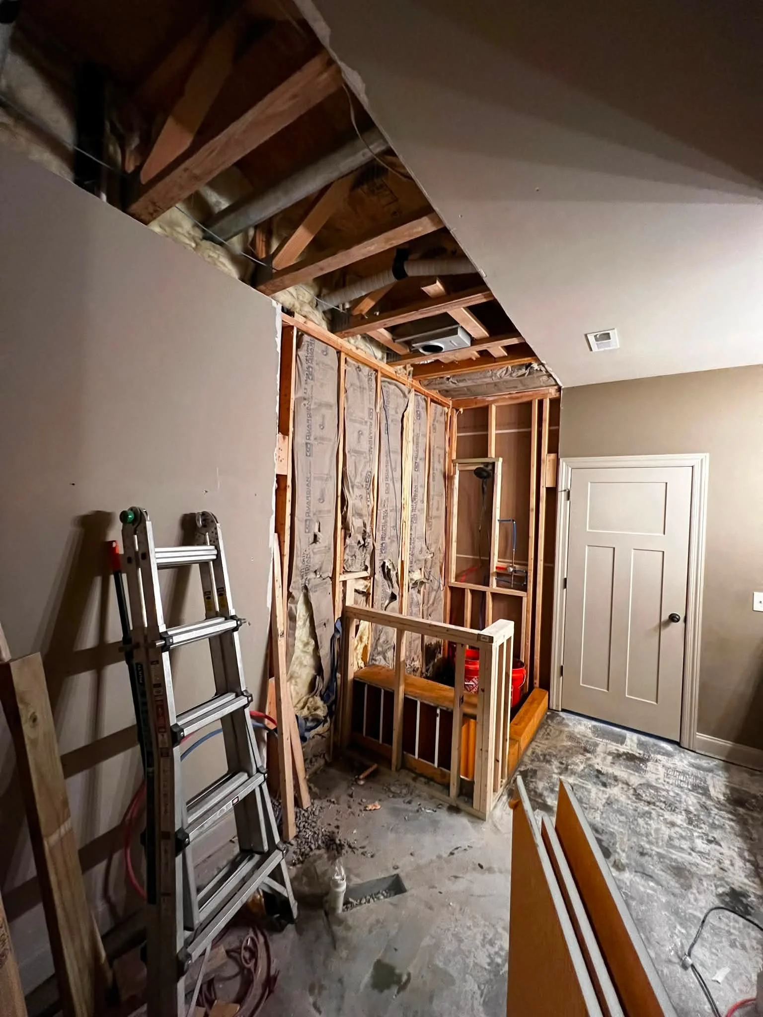 A room undergoing renovation with exposed wall studs, insulation, and pipes. There is a ladder leaning against the wall on the left side, and construction tools and materials scattered on the floor. Part of the ceiling is unfinished, and there's a do