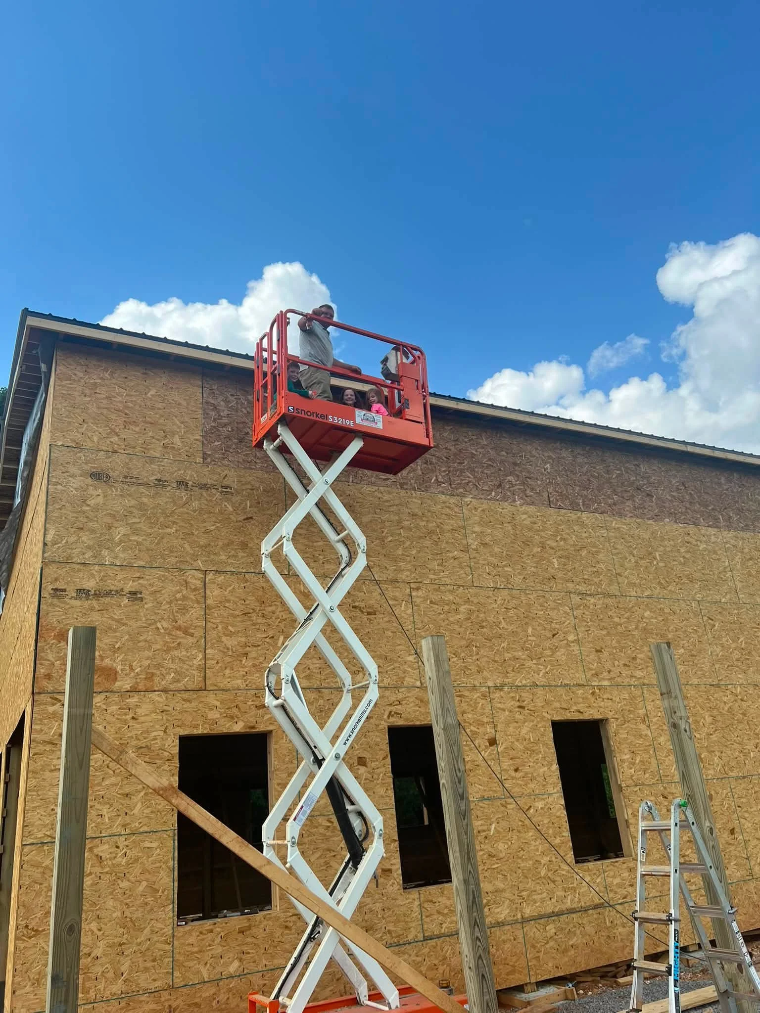 Three construction workers on an orange scissor lift working on the exterior of a building under construction. The building's exterior walls are covered with oriented strand board (OSB) sheathing, and there are window openings. The sky is mostly clea