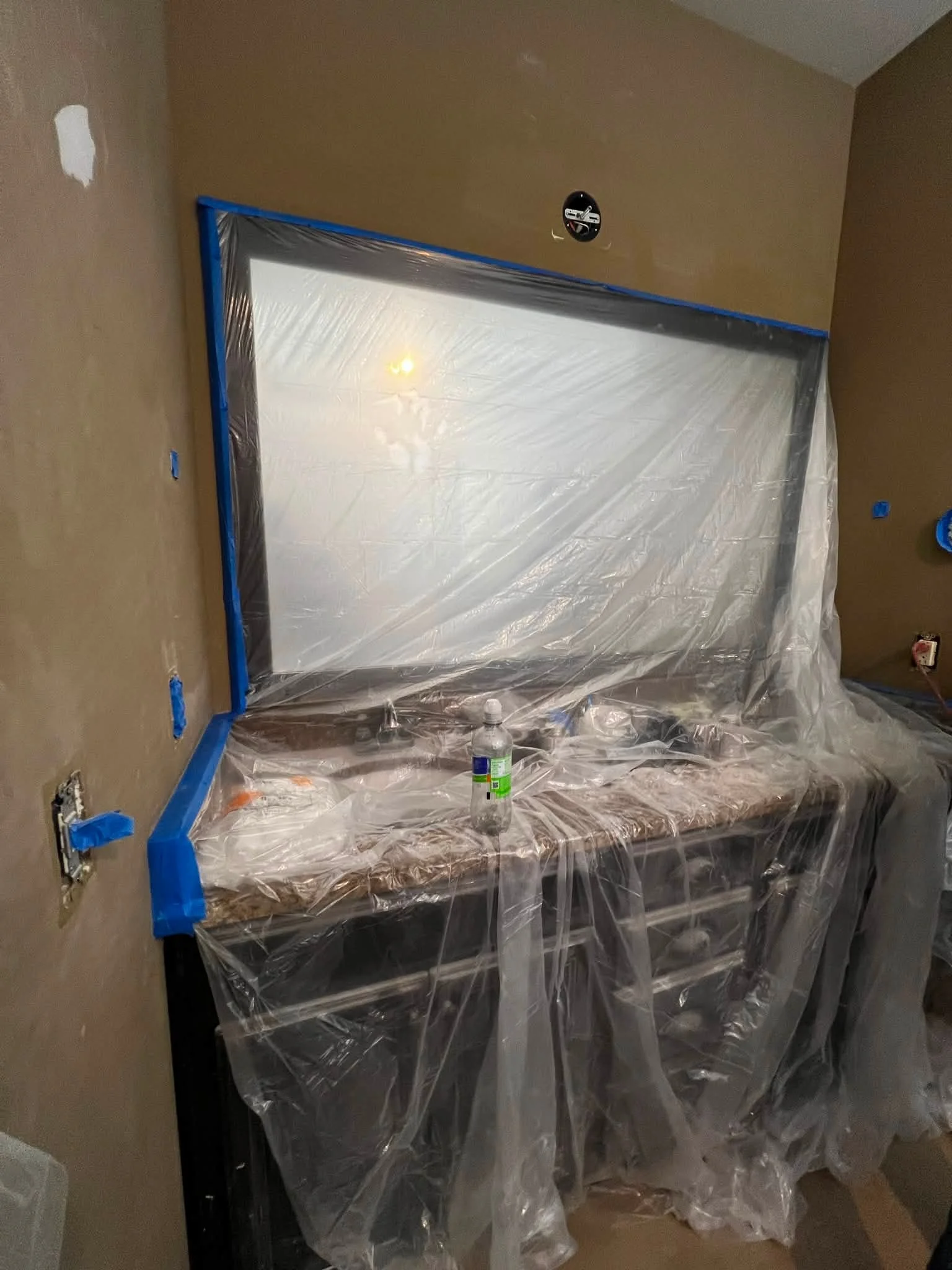 Room under renovation with a stove covered in plastic sheeting, taped edges, and surrounding walls with taped electrical outlets and switches, an exposed ceiling fixture, and a water bottle on the floor.