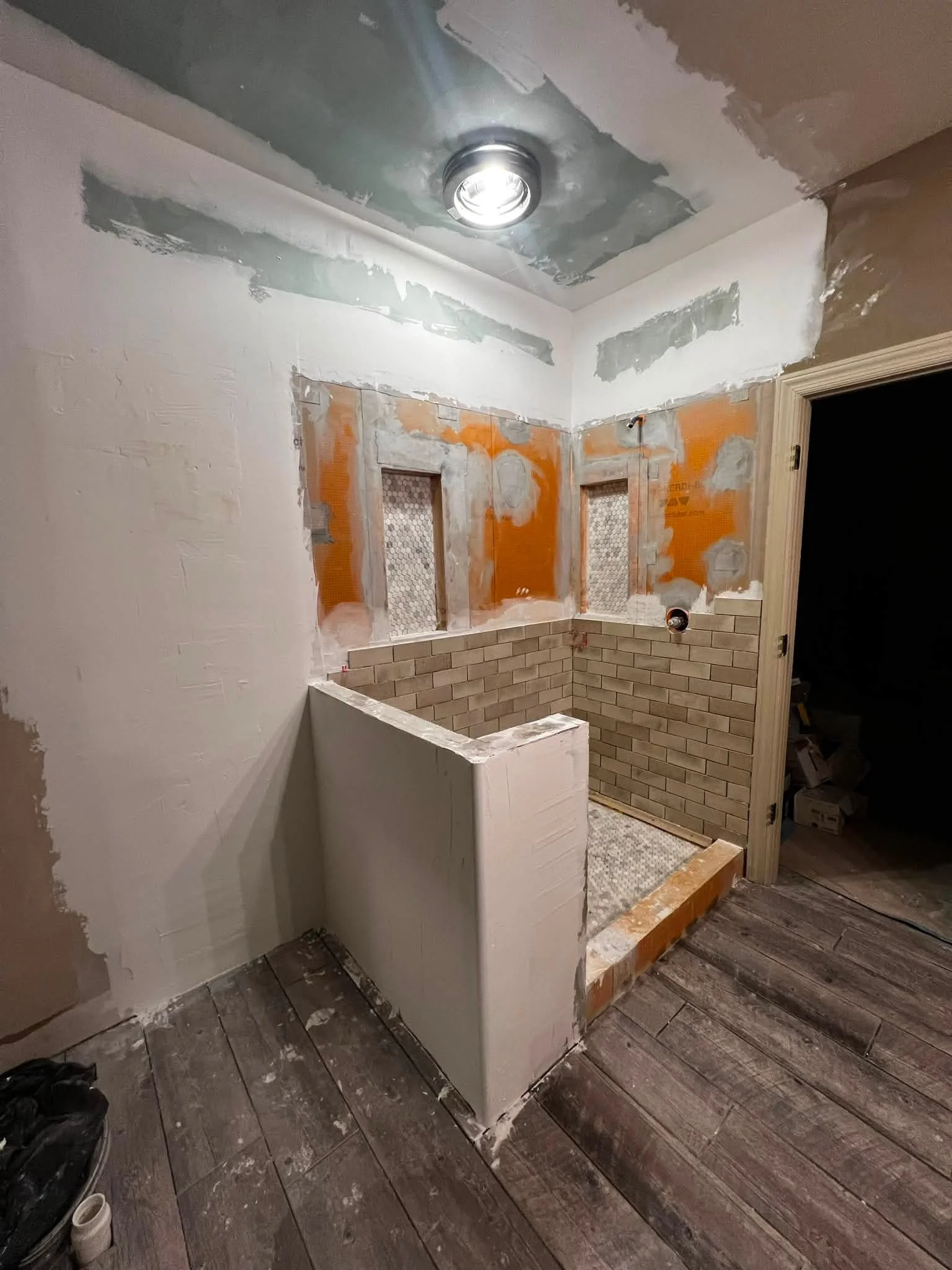 Bathroom under renovation showing drywall work, brick tiles on the lower wall, and a partially built shower stall with a white wall and a small section of custom tiling.