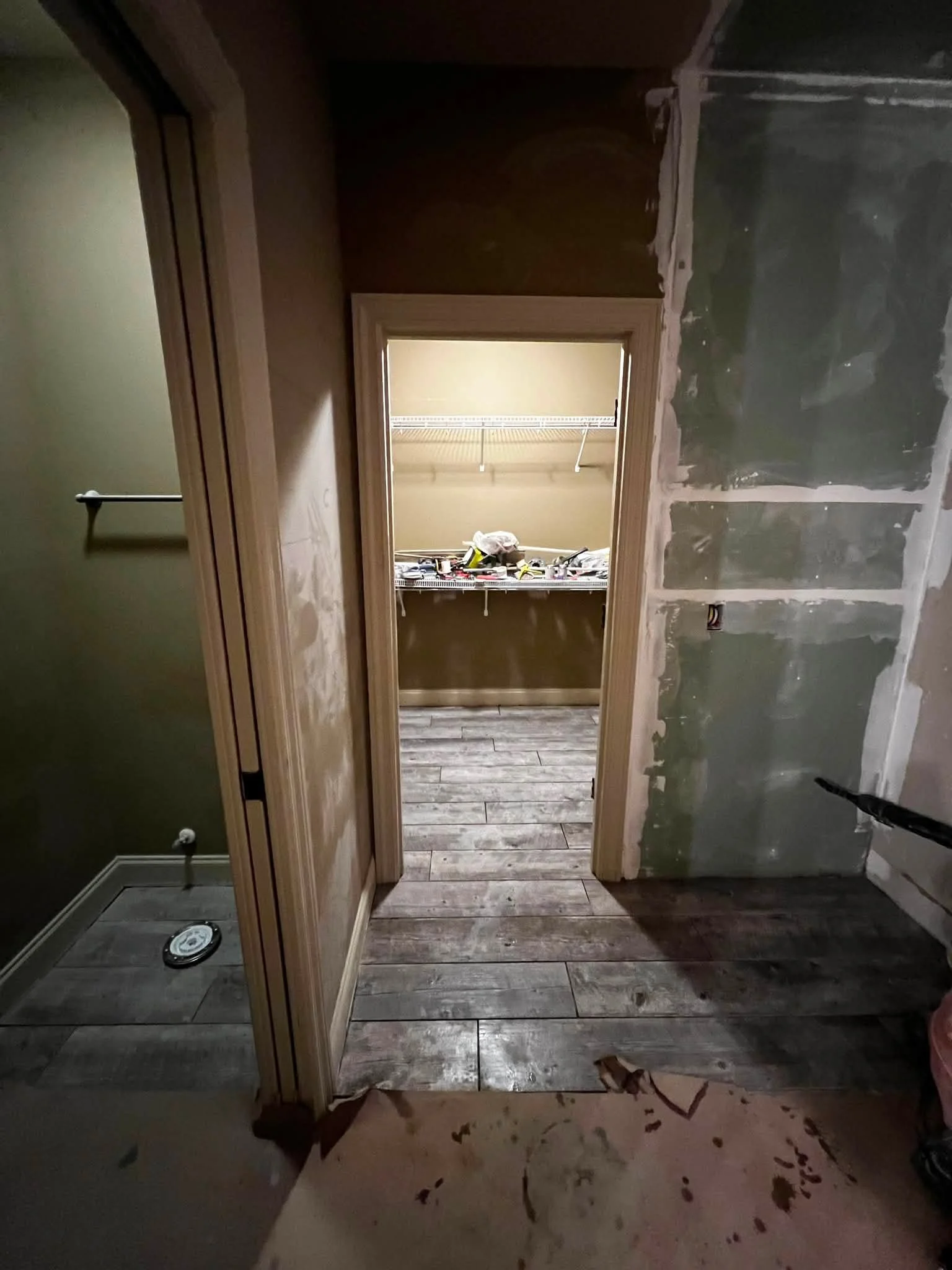 A room under construction or renovation with partially finished walls, a doorway with molding, and a wooden floor. Construction tools are on a shelf in the adjacent room, and the floor has debris.
