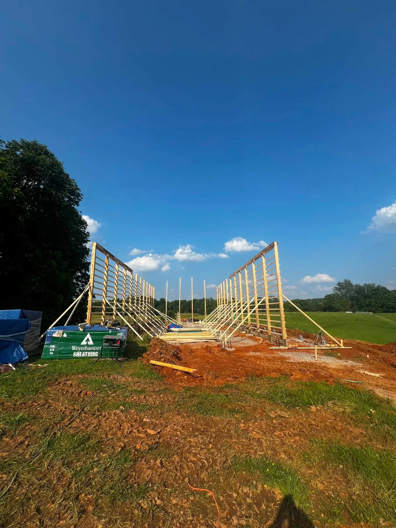 Construction site with a wooden frame structure in progress, on a grassy field with trees in the background. The sky is clear with some clouds.