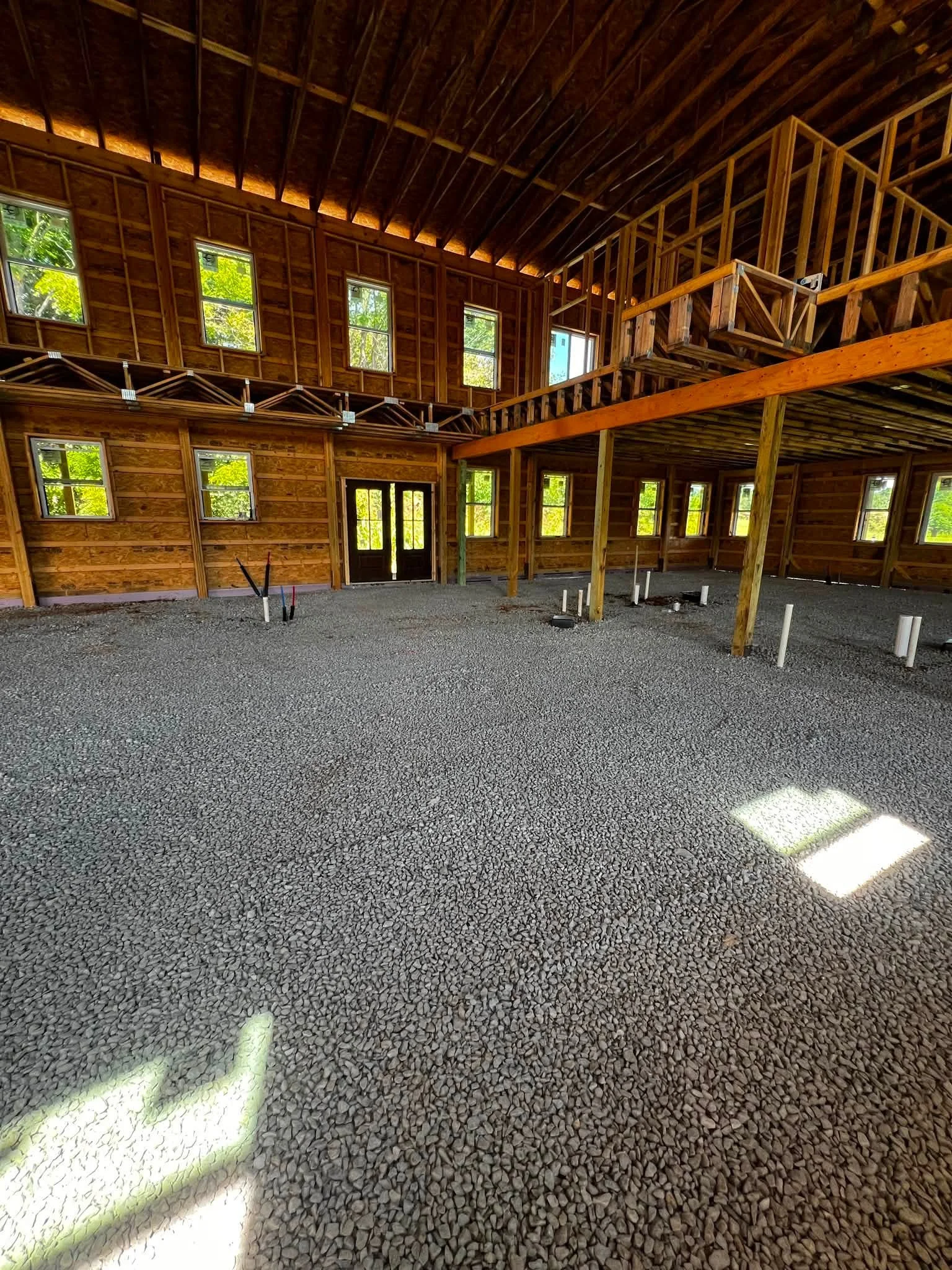 Interior of a wooden house under construction with exposed wooden beams, multiple windows, and a gravel floor.
