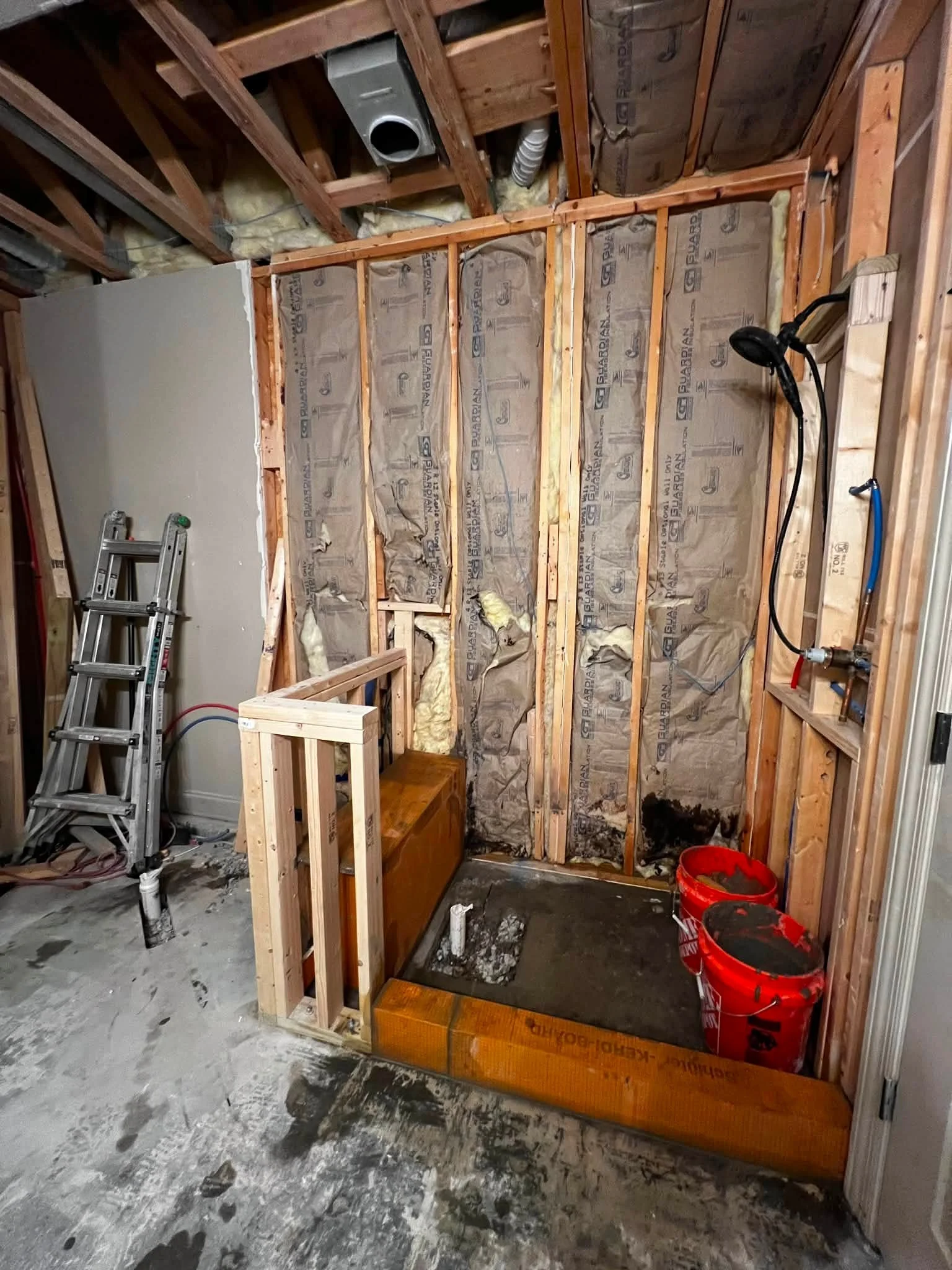 Construction site showing framed walls, insulation, and plumbing for a bathroom or utility room with tools, ladder, and buckets.