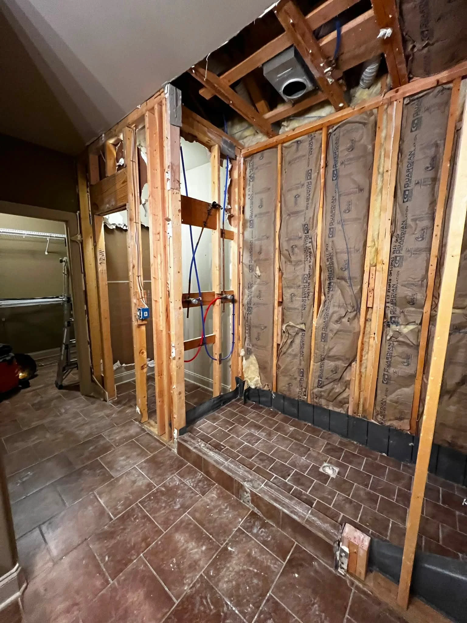 The photo shows a room under renovation with exposed wooden framing on the wall, insulation, and pipes. Part of the drywall is removed, revealing the interior structure.