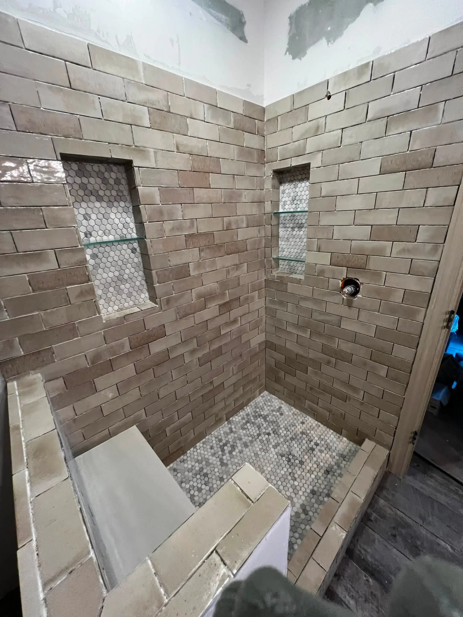 A bathroom shower area under construction with beige subway tiles on the walls, two inset niches with white hexagon tiles, and a mosaic tile floor in shades of gray and white.