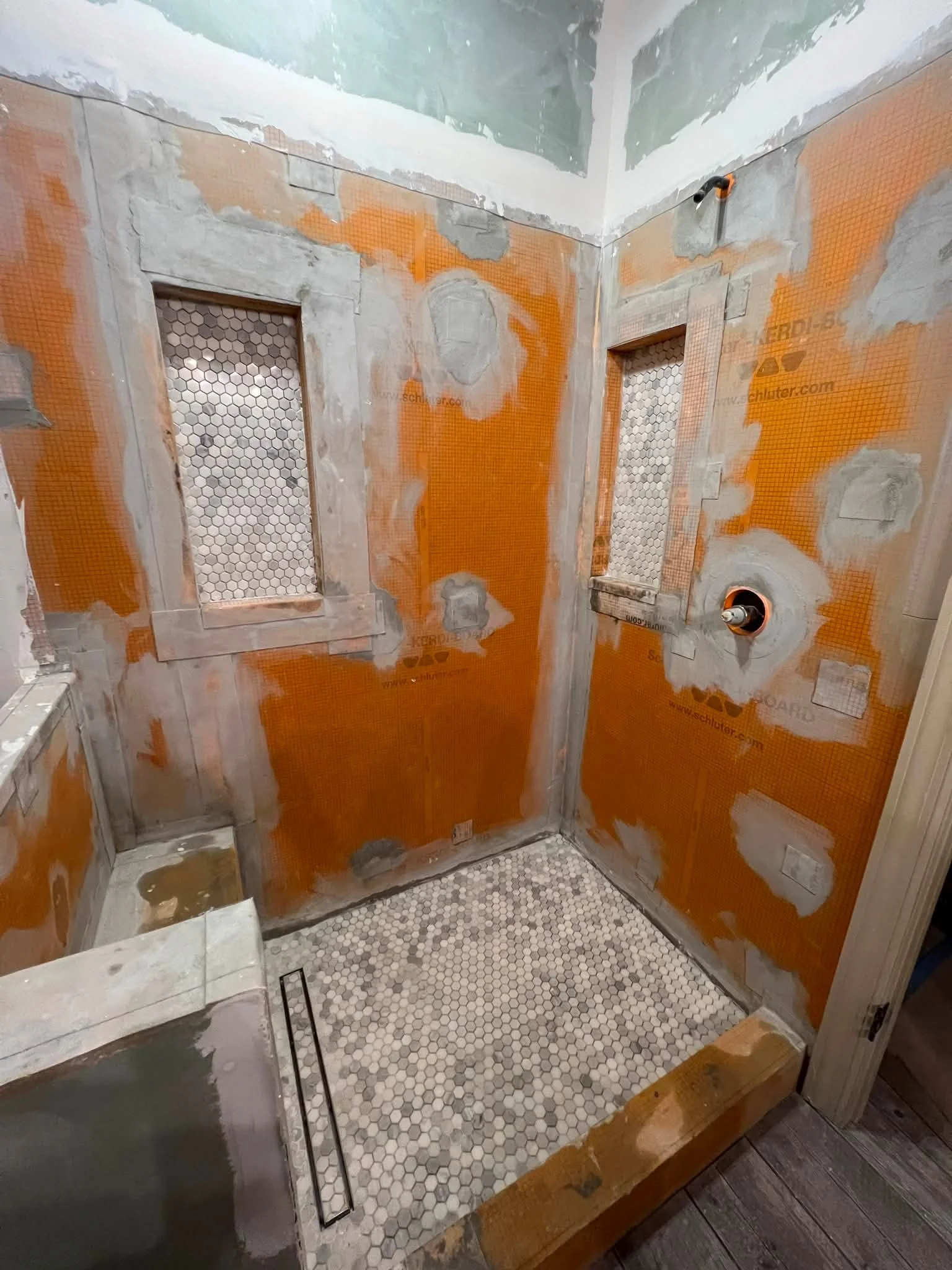 Bathroom under renovation with unfinished walls, two recessed niches with mosaic tile backs, and floors with small hexagon tiles.