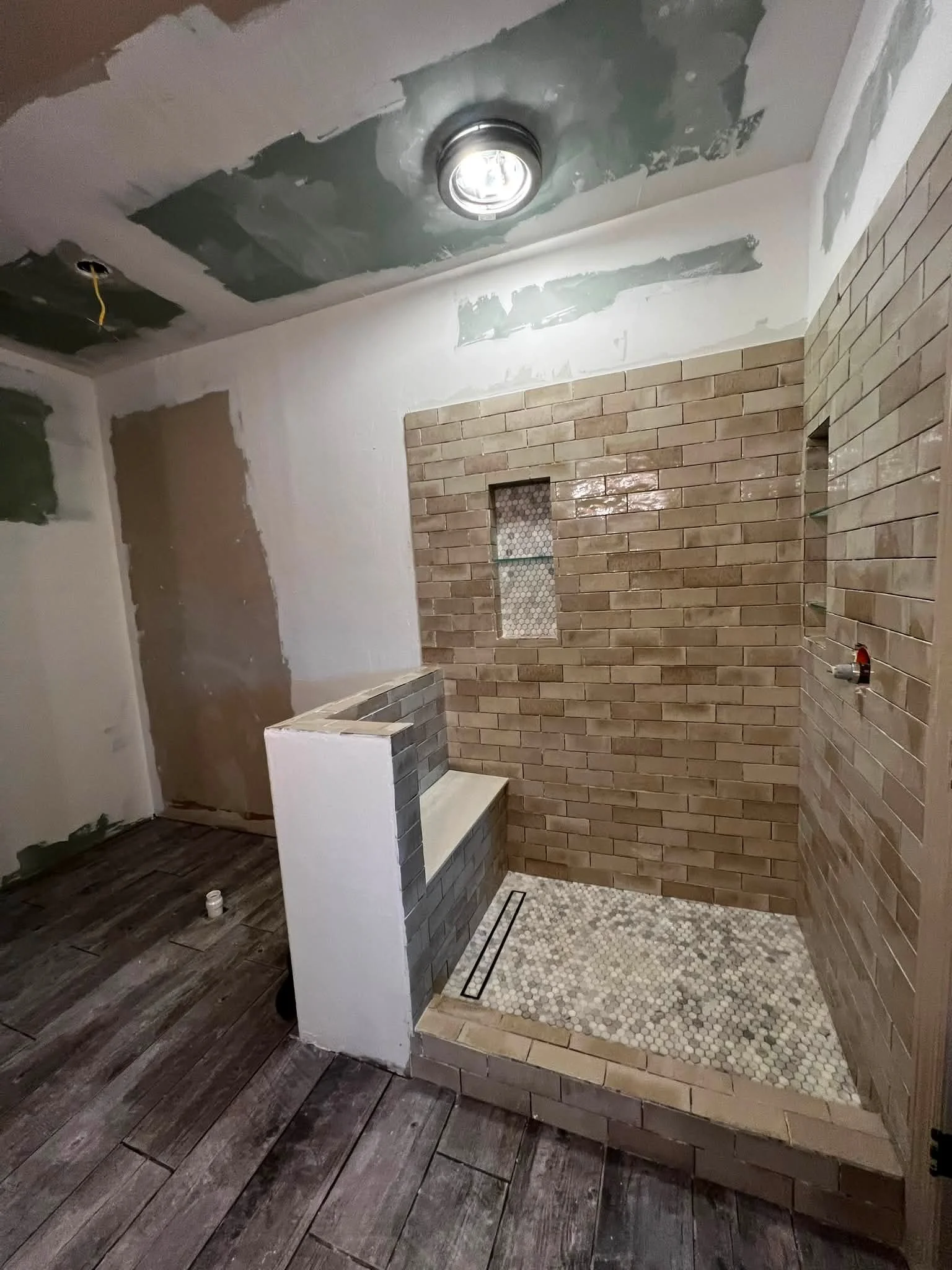 Bathroom under renovation showing a shower area with brick tiles, a small window, and stone flooring. The ceiling is unfinished with exposed drywall and a recessed light fixture.