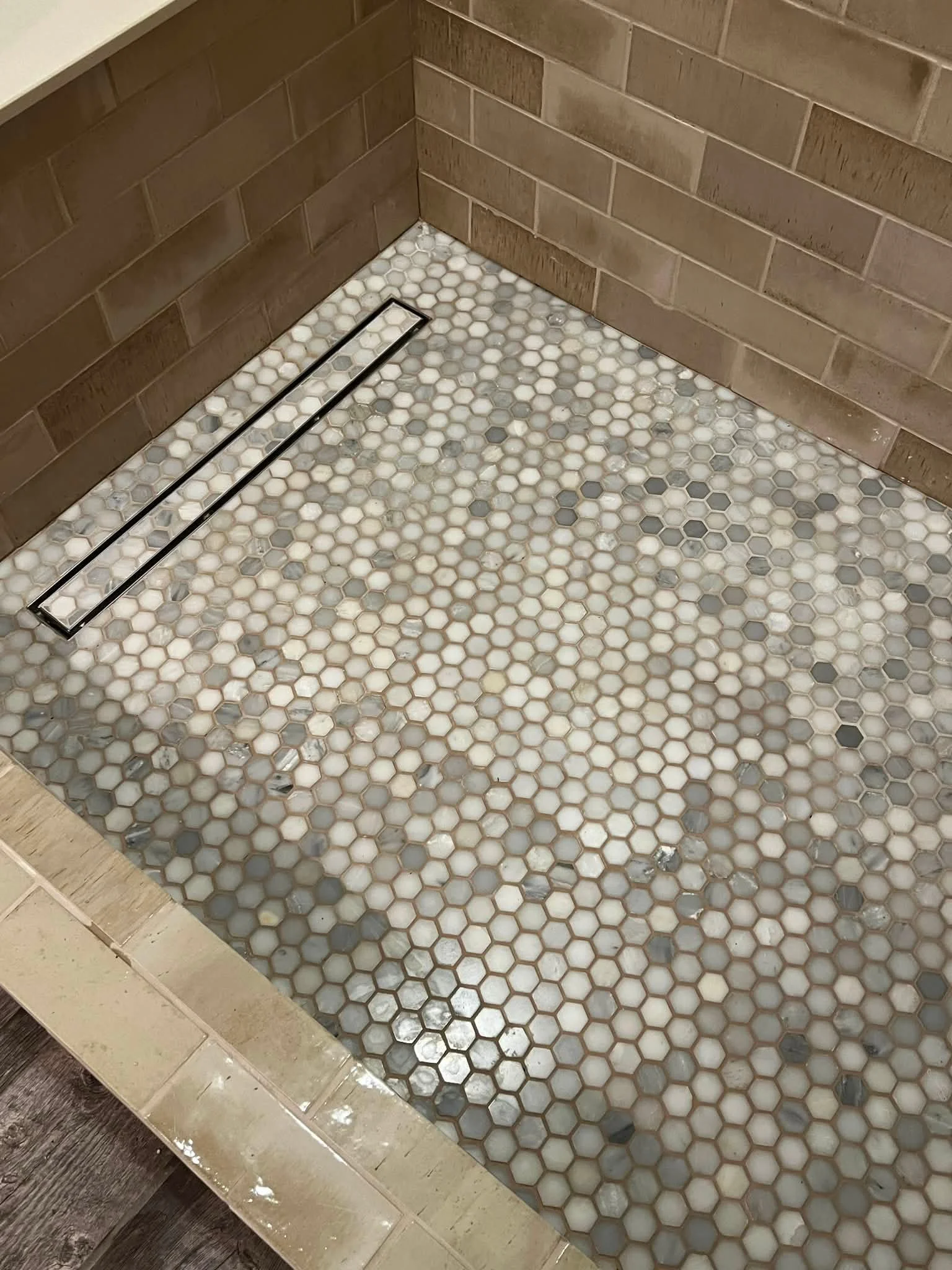 Bathroom floor with small hexagonal tiles in white, grey, and beige, a linear drain, and a wood-look tile border.