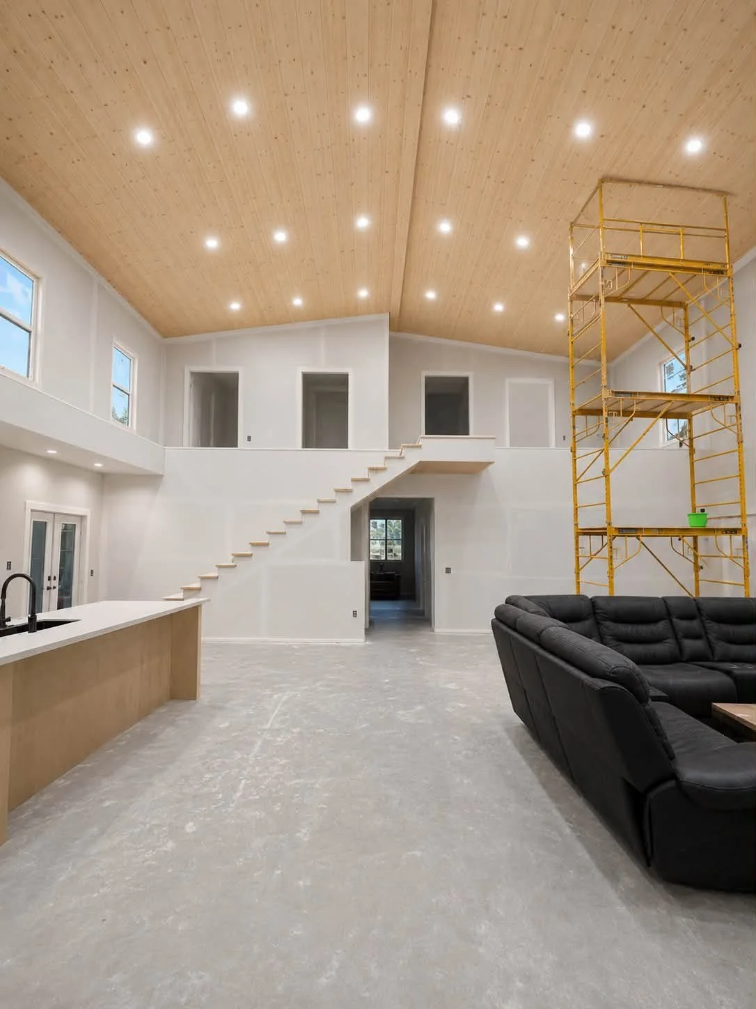 Interior of a modern house under construction with a high wooden ceiling, a black sofa, a kitchen island, a yellow scaffold, and white walls.