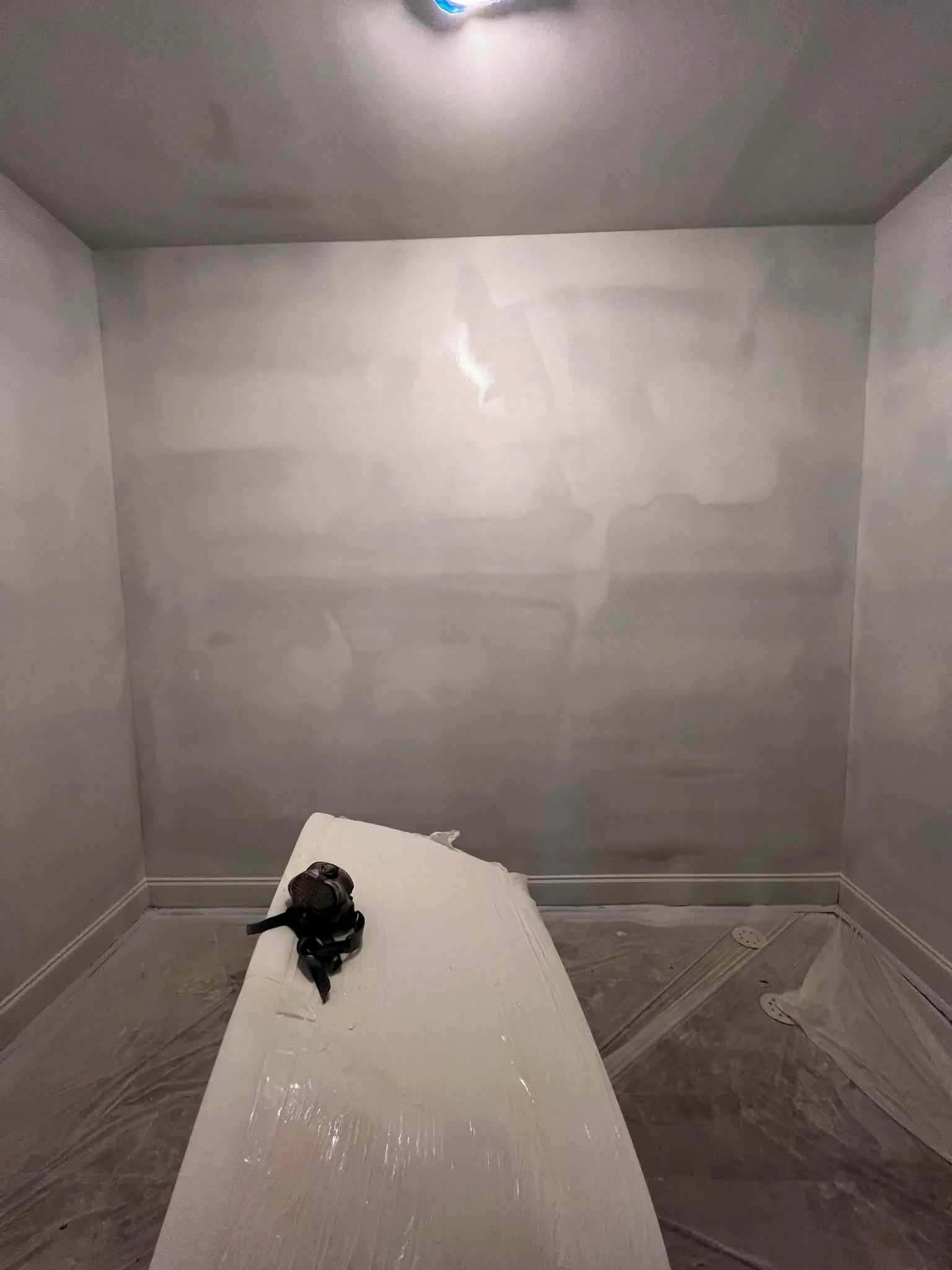 A room with grayish-white walls and ceiling, a light fixture on the ceiling, a piece of furniture covered with plastic on the floor, and a pair of black shoes resting on the furniture.