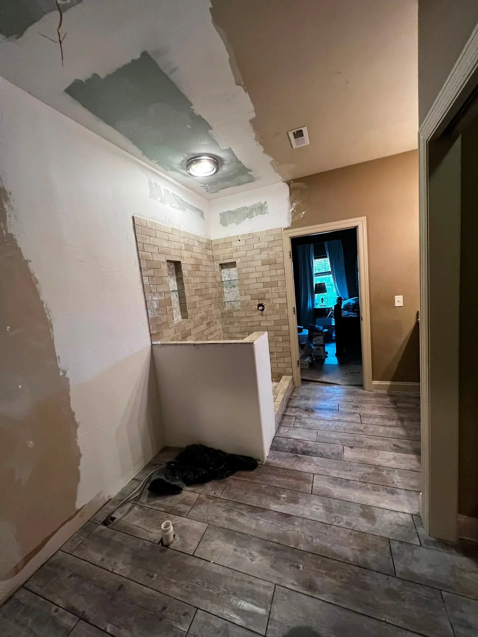 A room under renovation with partially finished walls and a ceiling with visible drywall patches. The floor has wood-like tiles, and there are construction tools and materials on the floor. An open doorway reveals a dimly lit room with a table lamp a