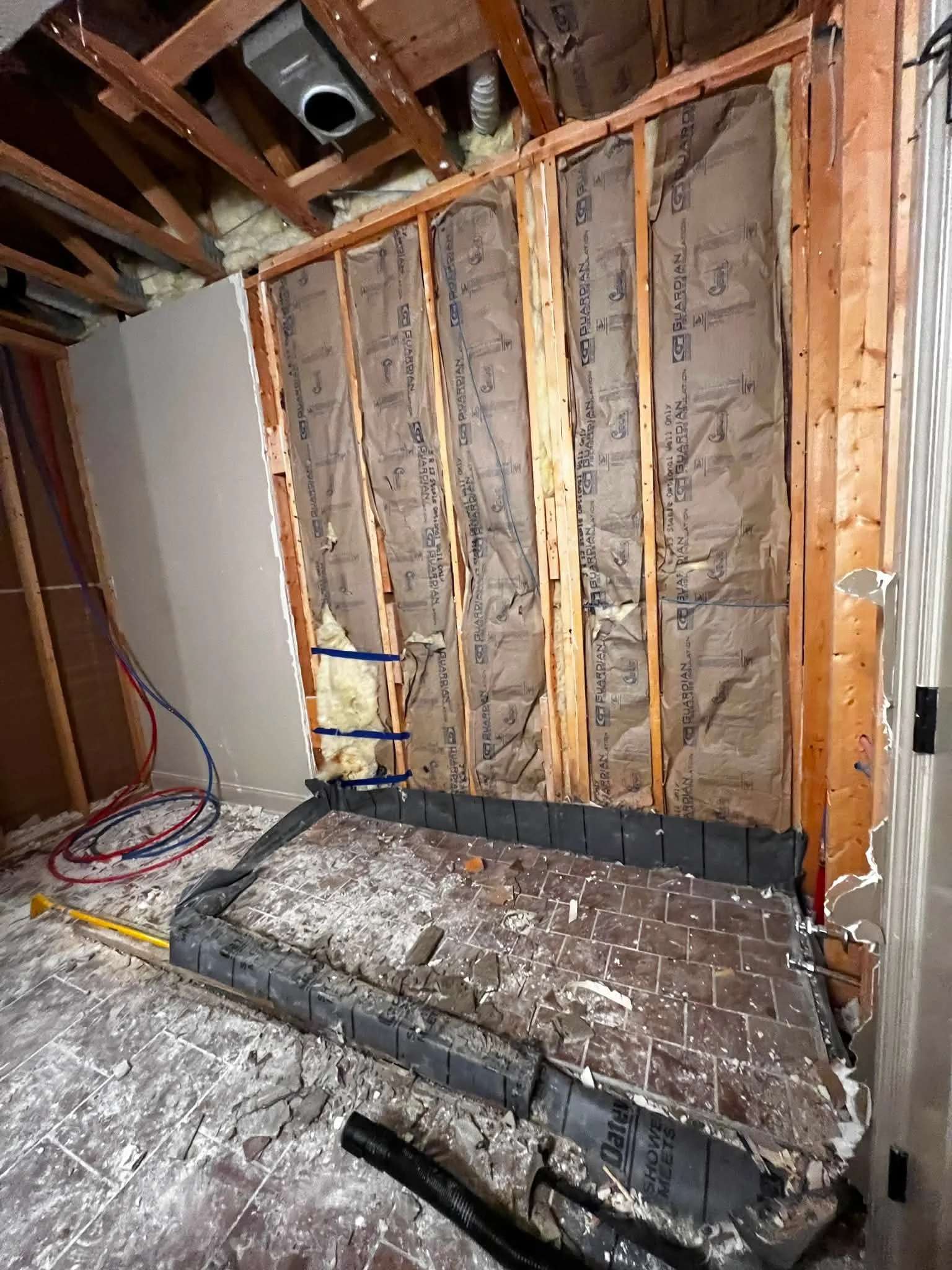 Construction or renovation scene with exposed wooden wall framing, insulation, and plumbing pipes. The floor is dirty with construction debris.